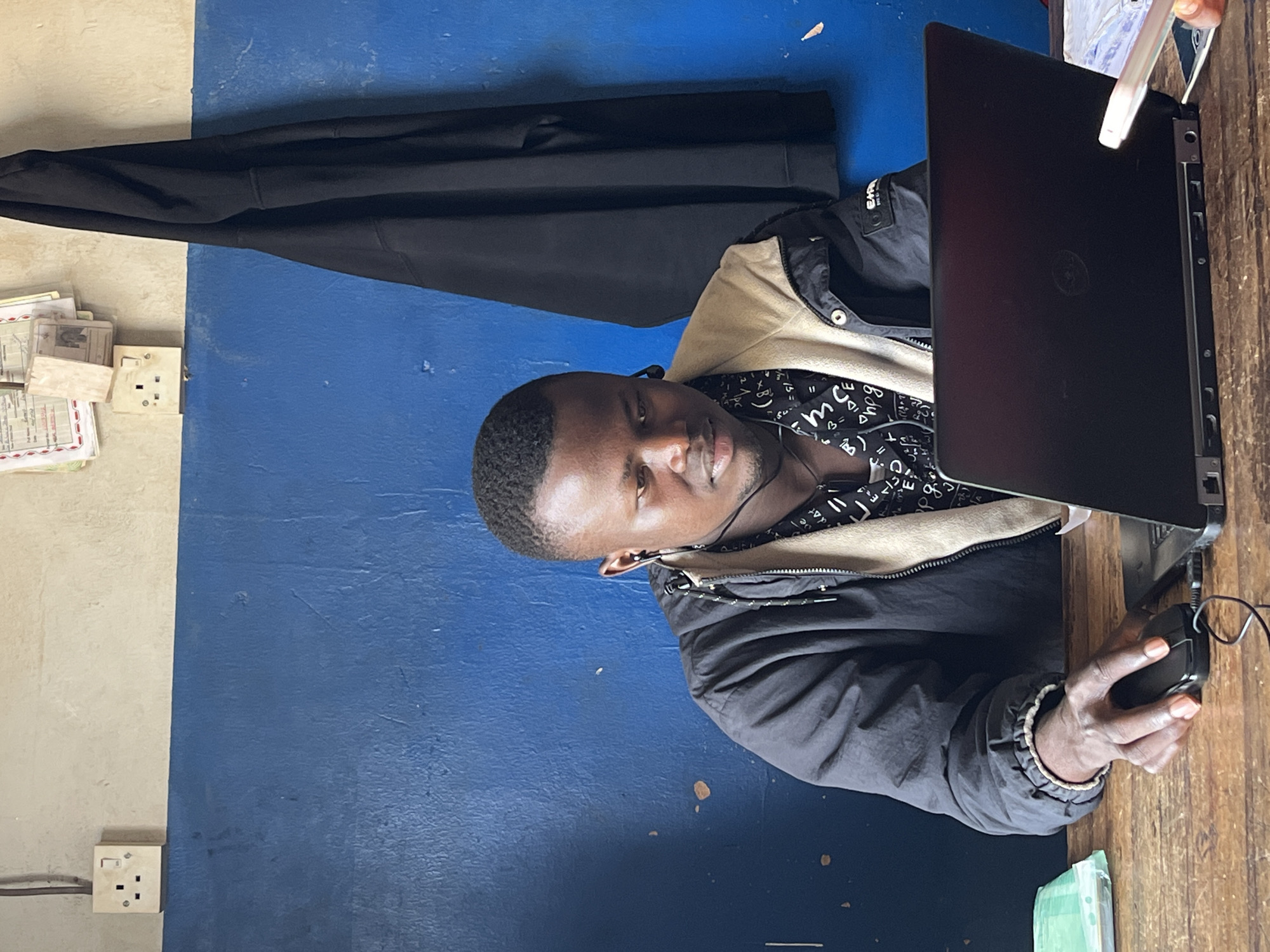 A young Black man with short hair and wearing earbuds is seated at a wooden desk in front of a laptop. He is wearing a black jacket over a black shirt with a white graphic print. His left hand is on a black computer mouse. The wall behind him is painted with a blue lower half and a cream upper half, with electrical outlets and papers visible on the cream section. A black garment, possibly a jacket or coat, is hanging on the wall. The scene appears to be an indoor office or workspace. The man's posture suggests he is actively engaged with the laptop. The lighting is moderate, illuminating the desk and the man.