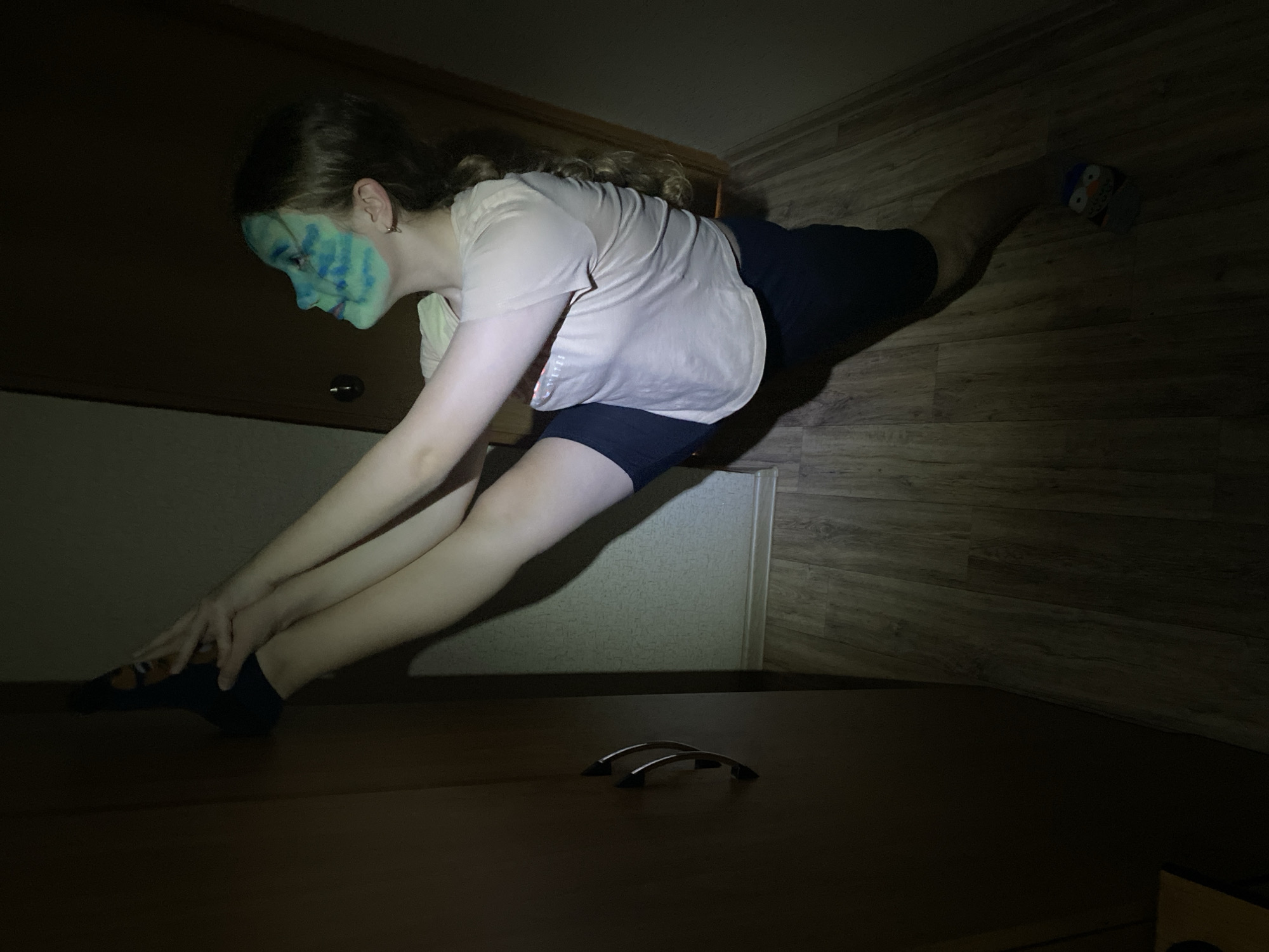 The image depicts a person with face paint, stretching in a dark room. The person is wearing a light-colored shirt and dark shorts. Their face has blue and green paint on it. They are stretching, reaching for their feet with their hands. The room has wood paneling on the walls and floor. There is a cabinet or drawer in the foreground with two metal handles. The lighting is dim, with a light source that appears to be focused on the person.