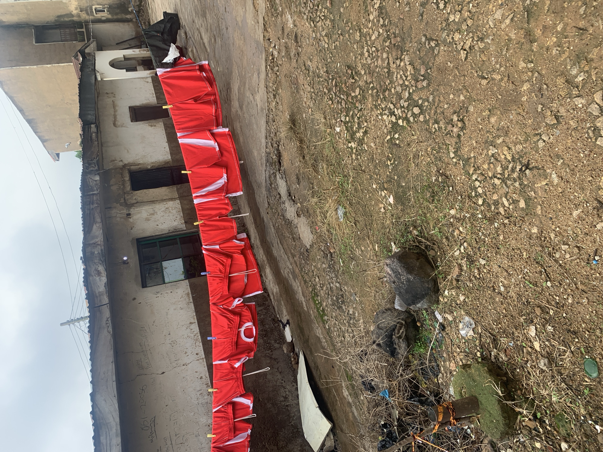 In the city of Unknown, Unknown, a weathered, dilapidated building with a corrugated metal roof is visible. The building's facade is stained and marked with graffiti. In front of the building, a clothesline strung with numerous red and white garments, likely shorts, hangs suspended. The garments are secured to the line with clothes pins. The ground in front of the building is rough and uneven, covered in dirt, rocks, and debris. Some dry grass and weeds are scattered across the area. To the left of the frame, a white, curved object, possibly a piece of salvaged material, lies on the ground. Further to the left, a metal post with orange bands is embedded in a moss-covered concrete base, surrounded by a pile of trash and branches. A dark colored bag and other refuse are also present. The overall scene suggests an outdoor area of a modest, possibly impoverished, urban setting.