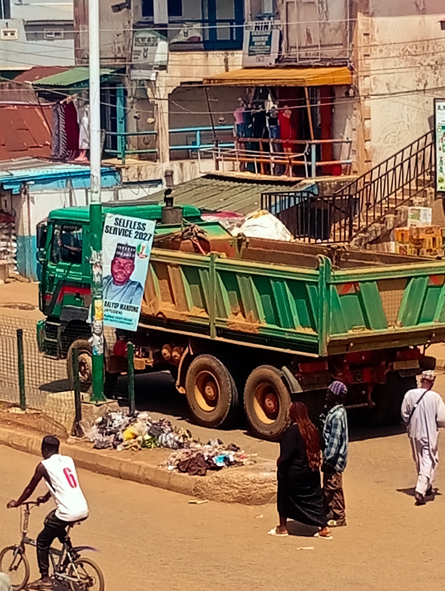 In Bukuru, Nigeria, a green dump truck is parked on a street with a campaign poster attached to a pole in front of it. The poster features a portrait of a man in traditional attire with the text "SELFLESS SERVICE 2027" and other inscriptions. A pile of refuse is visible on the roadside near the truck. To the left of the truck, a person is riding a bicycle. In the background, buildings with various colored roofs are visible, along with a staircase. To the right of the dump truck, three people are standing, and another person is walking away from the scene. The environment appears to be an urban street with some commercial activity suggested by the storefronts and awnings.