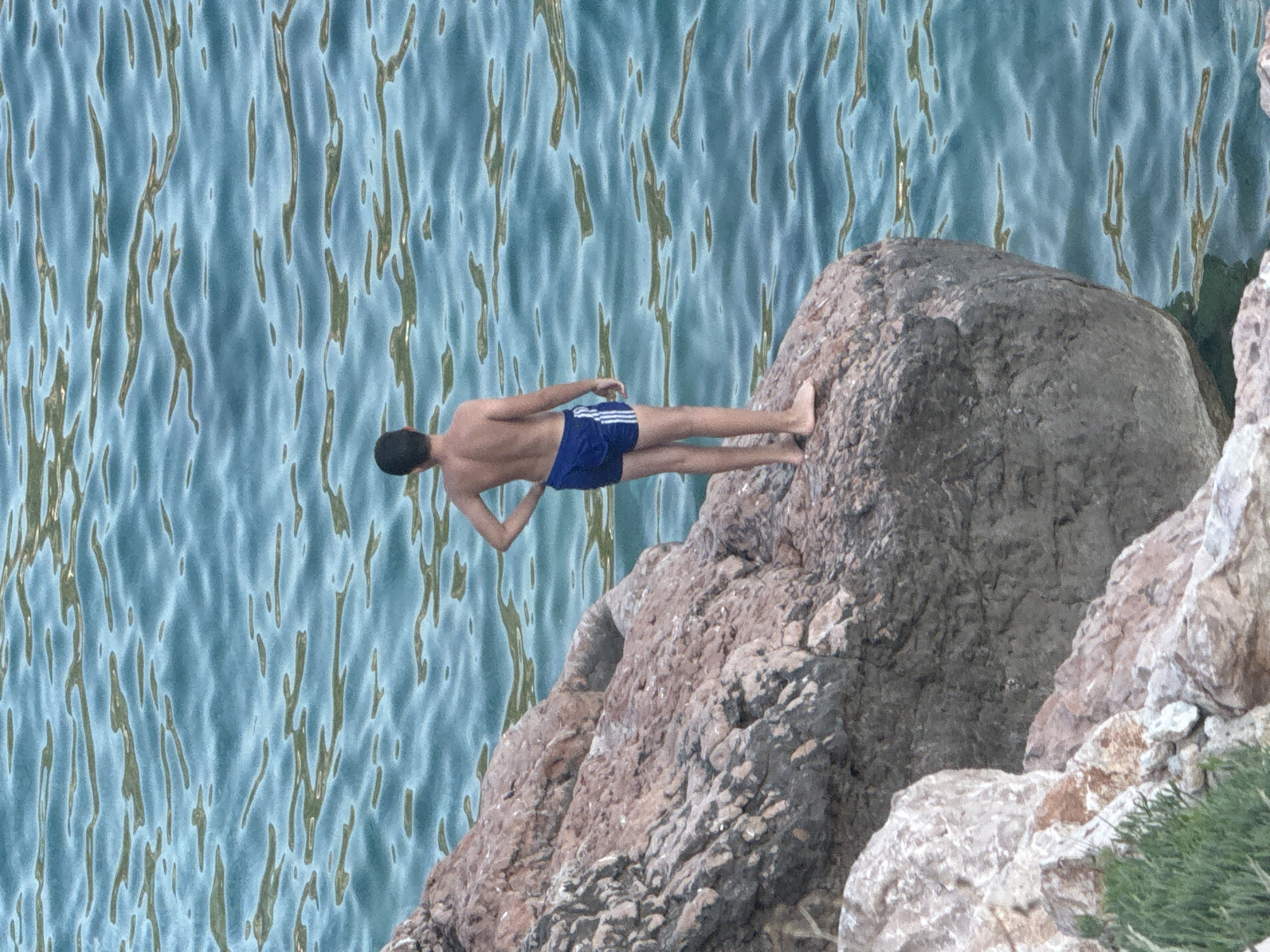 A young male, from the back, stands on a rocky outcropping in Kamenovo, Montenegro. He is wearing blue swim trunks with white stripes. The water is a clear blue with visible ripples. The rocky terrain is rough and textured. Green foliage is visible at the bottom left of the image.