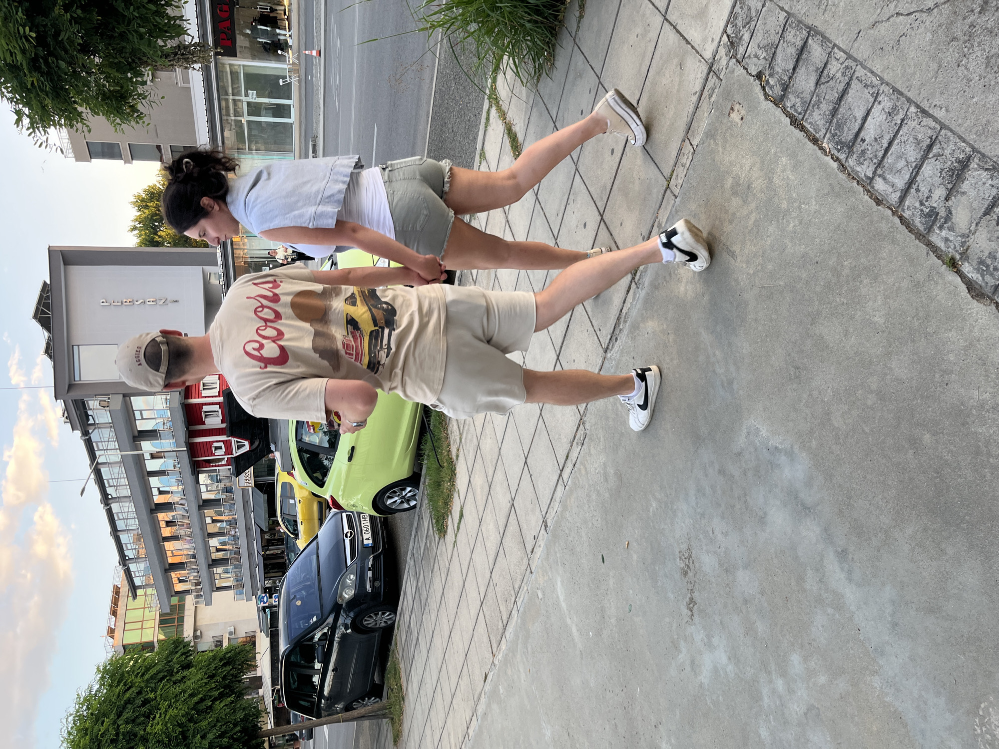 Two individuals, a male and a female, are walking hand-in-hand along a paved sidewalk. The male, facing away from the camera, wears a light-colored t-shirt with a graphic print and light-colored shorts, and white sneakers with black accents. The female, also facing away from the camera, wears a white tank top, light-colored denim shorts, and platform sneakers.
They are walking on a sidewalk in front of a row of buildings, indicating an urban or town setting. Behind them, cars are parked along the street. A dark-colored minivan is parked on the left, and a bright green compact car is to its right. Further down the street, a yellow taxi is visible. The buildings appear to be commercial establishments, with windows and signage. One building has a distinctive red facade with white accents. The sky above is partly cloudy with a warm, late afternoon or early evening light. The overall scene suggests a casual stroll through a developed area, likely in a tourist destination given the variety of vehicles and commercial fronts. The location is identified as the city of Nesebar, Bulgaria.