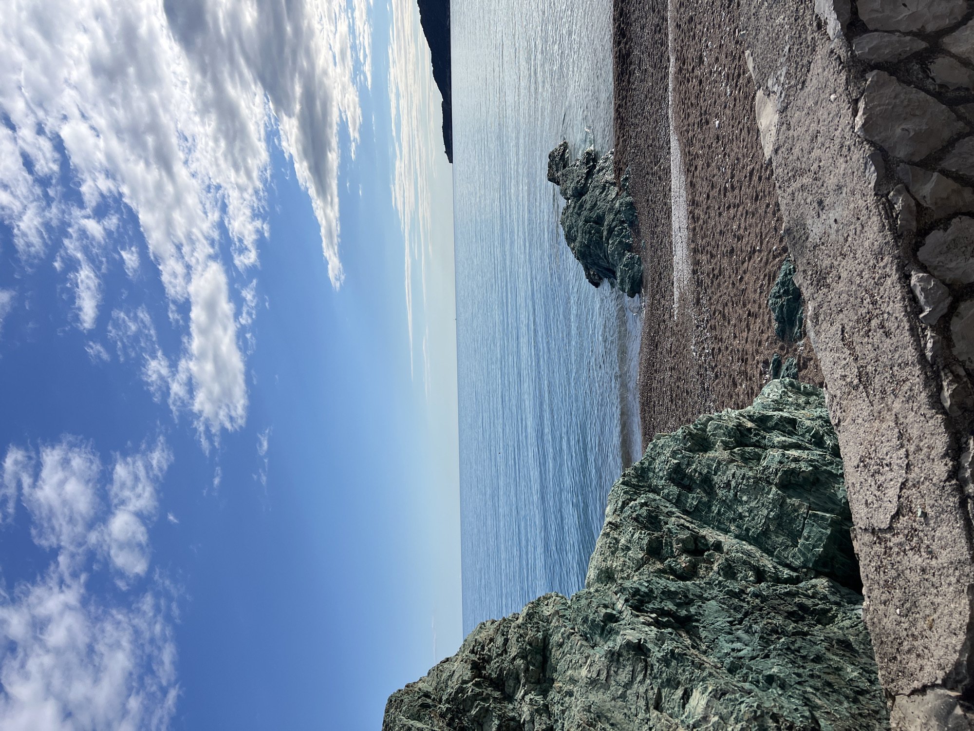 The image depicts a coastal view in Rafailovići, Montenegro. The composition is divided vertically, with the left side featuring a blue sky with scattered white clouds. The right side shows the Adriatic Sea extending to a distant, dark landmass. A rocky beach is visible between the sea and a partially visible stone wall. The foreground contains a large, green-tinged rock formation.