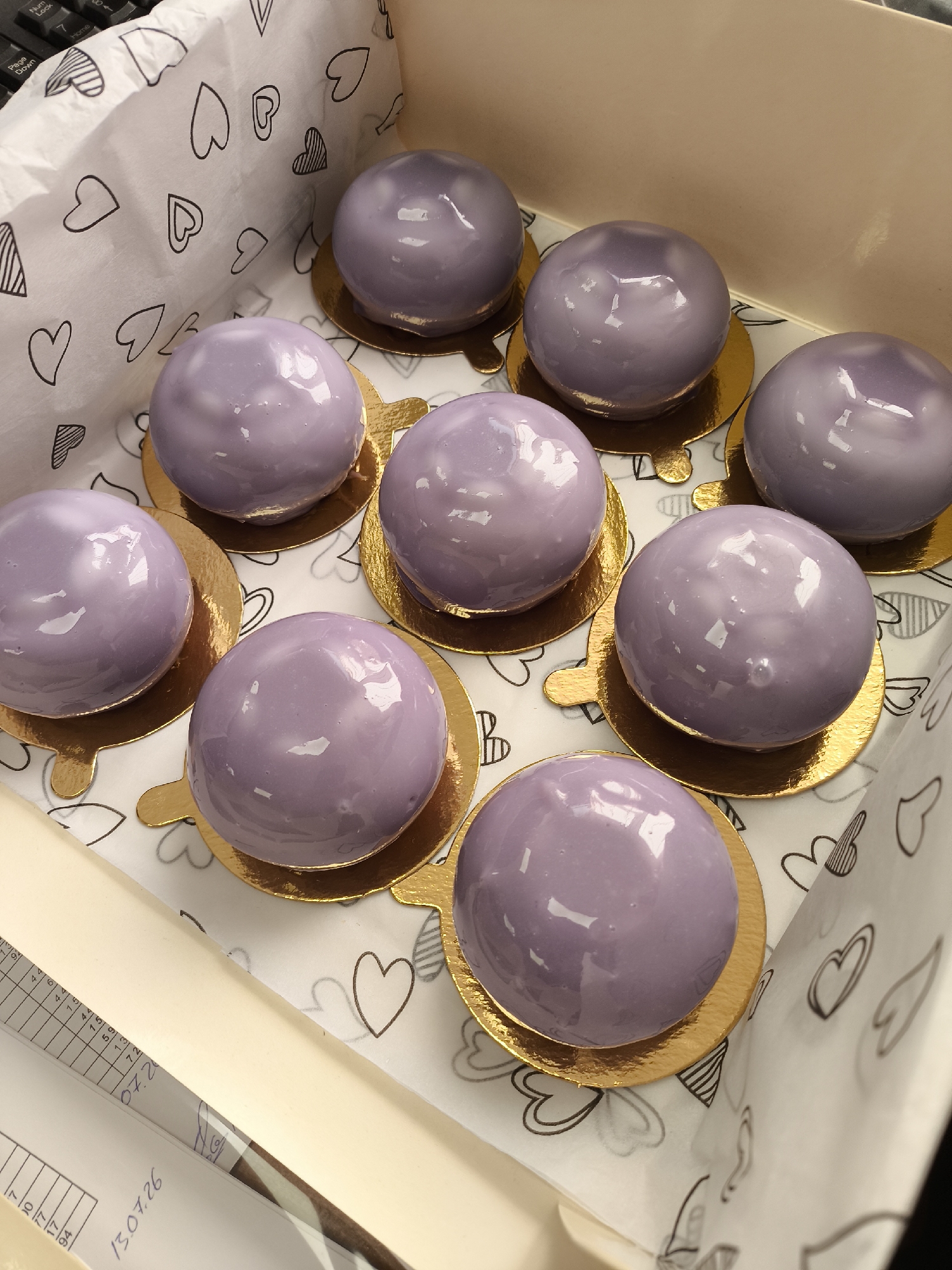 A high-angle medium shot captures a box of nine individual, spherical, purple glazed pastries. The pastries are arranged in three rows of three. Each pastry rests on a gold circular base. The interior of the box is lined with white paper featuring a repeating pattern of black outline hearts of various sizes and styles. The pastries are evenly spaced within the box, with the top center pastry slightly more in focus than the others. In the bottom left corner of the image, a portion of a document with handwritten numbers and text is visible. The background of the box is a light tan color.
