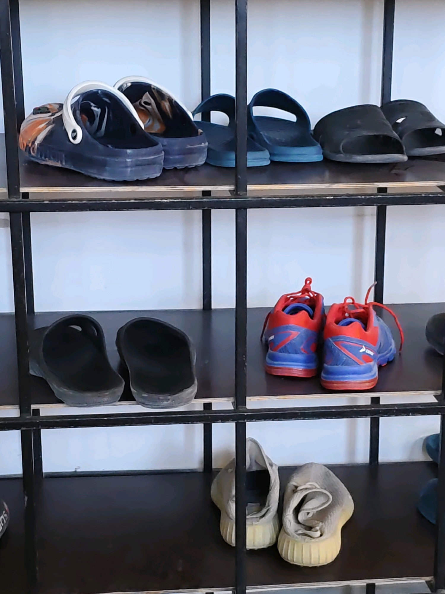 The image displays a black metal shelf unit with several pairs of shoes arranged on its shelves. The shelves are dark brown.
On the top shelf, there are two pairs of dark blue clogs with white straps. To their right are two pairs of dark blue slide sandals. Further right are two black slides.
The second shelf from the top features two black clogs on the left side. On the right side of this shelf are two blue and red athletic shoes with laces.
The third shelf from the top is mostly empty, with only a glimpse of a dark shoe on the far right.
The bottom shelf has a pair of light grey slip-on sneakers on the left, and further to the right, a dark blue slide sandal is visible. The background appears to be a plain white wall.
The scene is an indoor setting, likely a storage area or a room in a residence. There are no people visible in the image. The shoes are the primary focus, suggesting a collection or storage of footwear. The location is stated to be Rostov-on-Don, Russia.