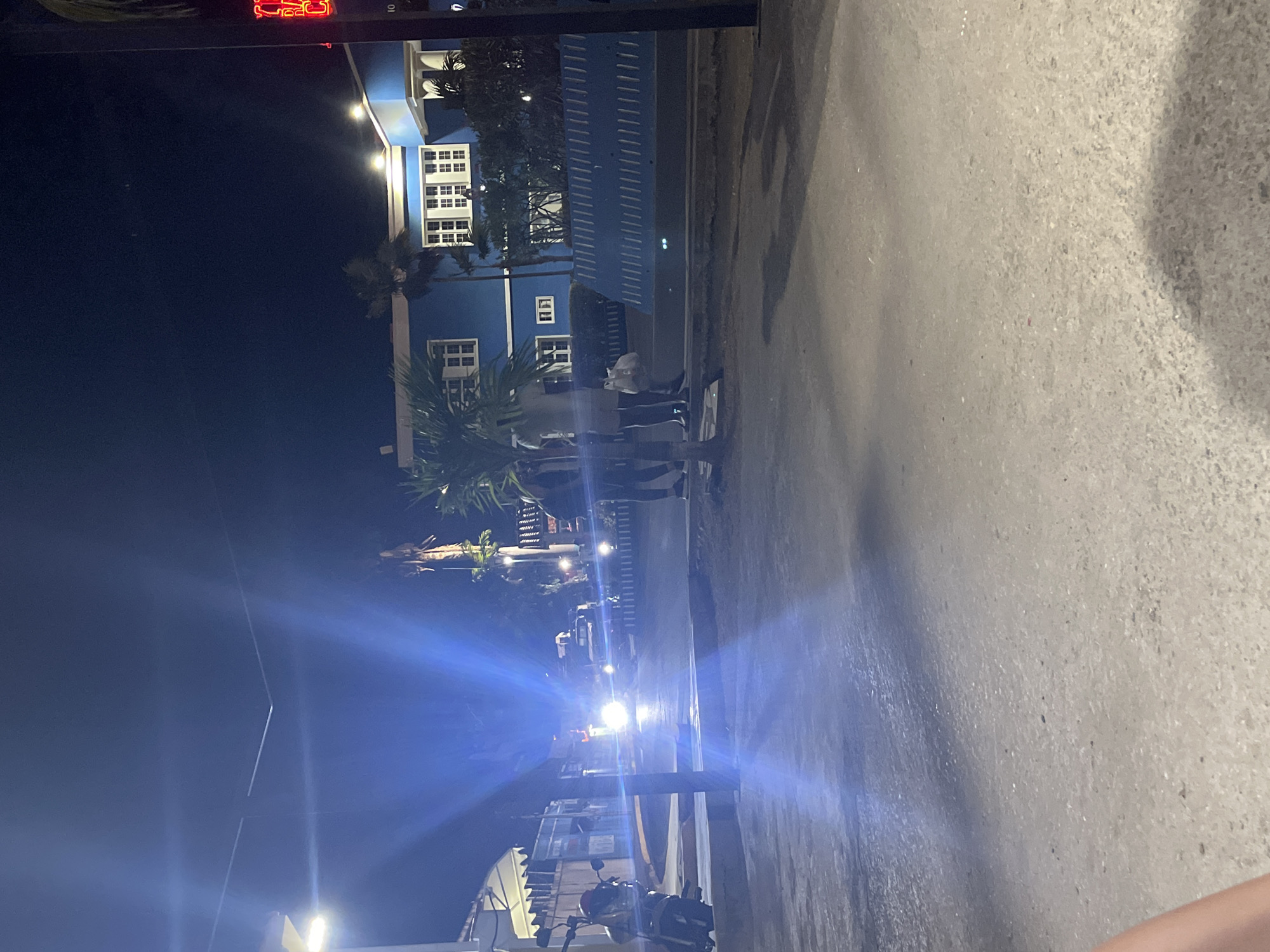 At night in Juan Griego, Venezuela, a street scene shows a blue building with multiple windows and a red neon sign near the top, facing the road. The building is partially obscured by palm trees. Several people are standing on the sidewalk near the building, some wearing light-colored clothing. Cars with headlights on are traveling along the street, their lights creating lens flares. A parked motorcycle is visible in the foreground near the bottom of the frame. The road appears wet and reflects some of the light.