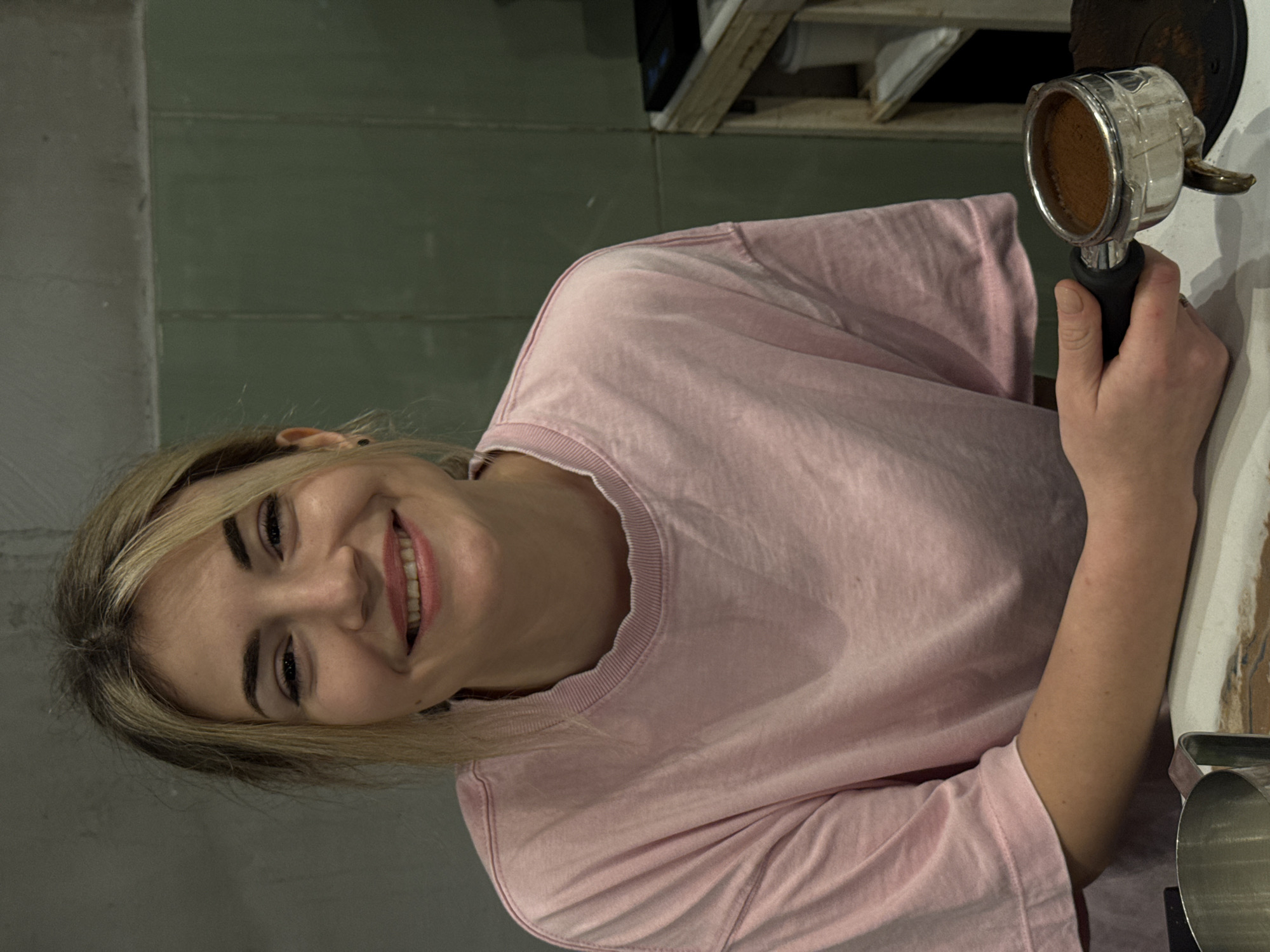 A young Caucasian female with blonde hair and bangs smiles directly at the viewer, positioned in what appears to be a coffee preparation area within a facility in Budva, Montenegro. She wears a loose-fitting, light pink, long-sleeved crew-neck shirt. Her left hand holds a silver metal portafilter filled with finely ground coffee, resting on a dark, circular mat on a light-colored countertop. A stainless steel milk frothing pitcher is partially visible to her left on the same counter. The background consists of a textured gray wall to her left and a dark green, paneled wall to her right, behind which a wooden shelving unit with miscellaneous items, including a black electronic device and white cups, is partially visible.