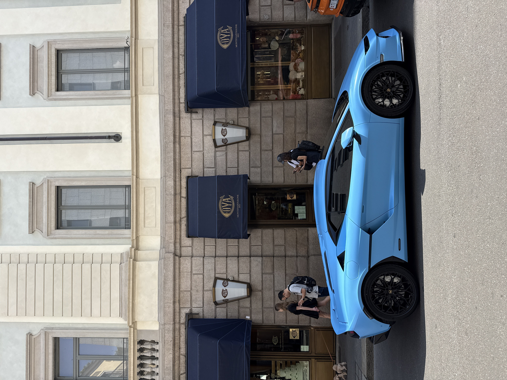 A turquoise Lamborghini sports car is parked on the side of a street in front of a building. The building has three dark blue awnings with the name "OVA Montenapoleone 1917" printed on them. There are two windows on the upper levels of the building. A couple is standing on the sidewalk in front of the building. A man with a backpack is walking past them. The location is Milan, Italy. An orange vehicle is partly visible at the top of the frame.