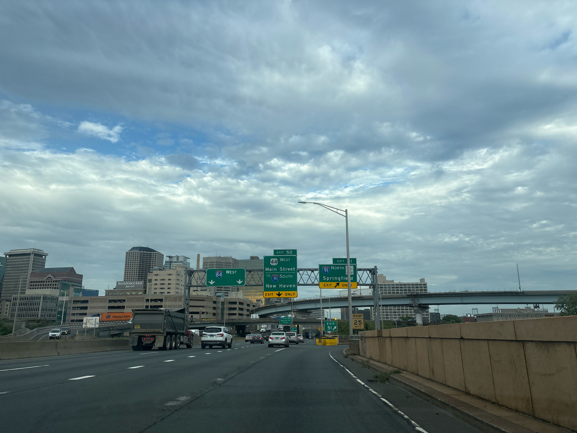 This image captures a highway scene in Hartford, Connecticut, USA. The view is from inside a vehicle driving on a multi-lane highway under an overcast sky with dramatic clouds. Road signs indicate exits for various destinations including "West 84", "Main Street", "New Haven", and "Springfield". A large dump truck is prominently featured in the foreground, alongside other cars and SUVs. In the background, a city skyline is visible with several tall buildings, including a "Residence Inn Marriott". Elevated highway ramps and bridges crisscross the scene, suggesting a complex urban interchange. A concrete barrier lines the right side of the highway, with some vegetation growing along its base. A speed limit sign of "25 MPH" is visible on a ramp on the right.