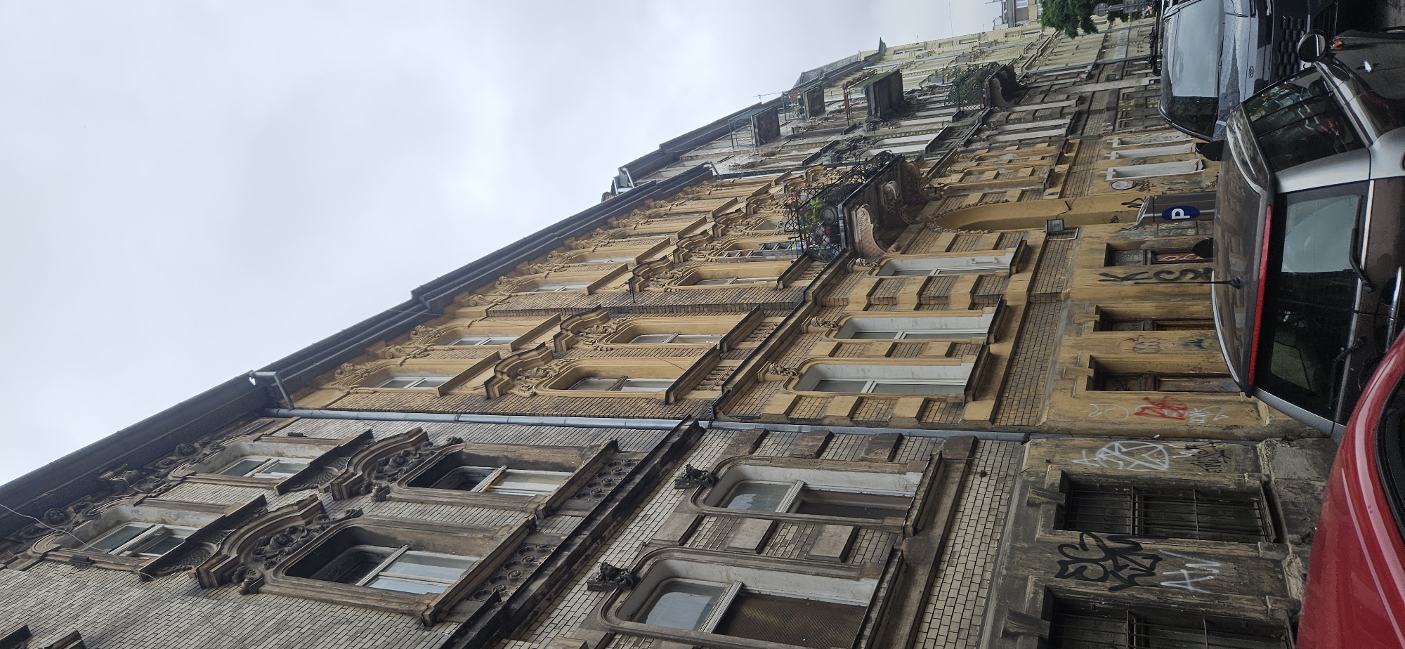 The image shows a street scene in Krakow, Poland. The buildings are old, with ornate facades and balconies. There are cars parked on the street, and graffiti on some of the walls. The sky is overcast.