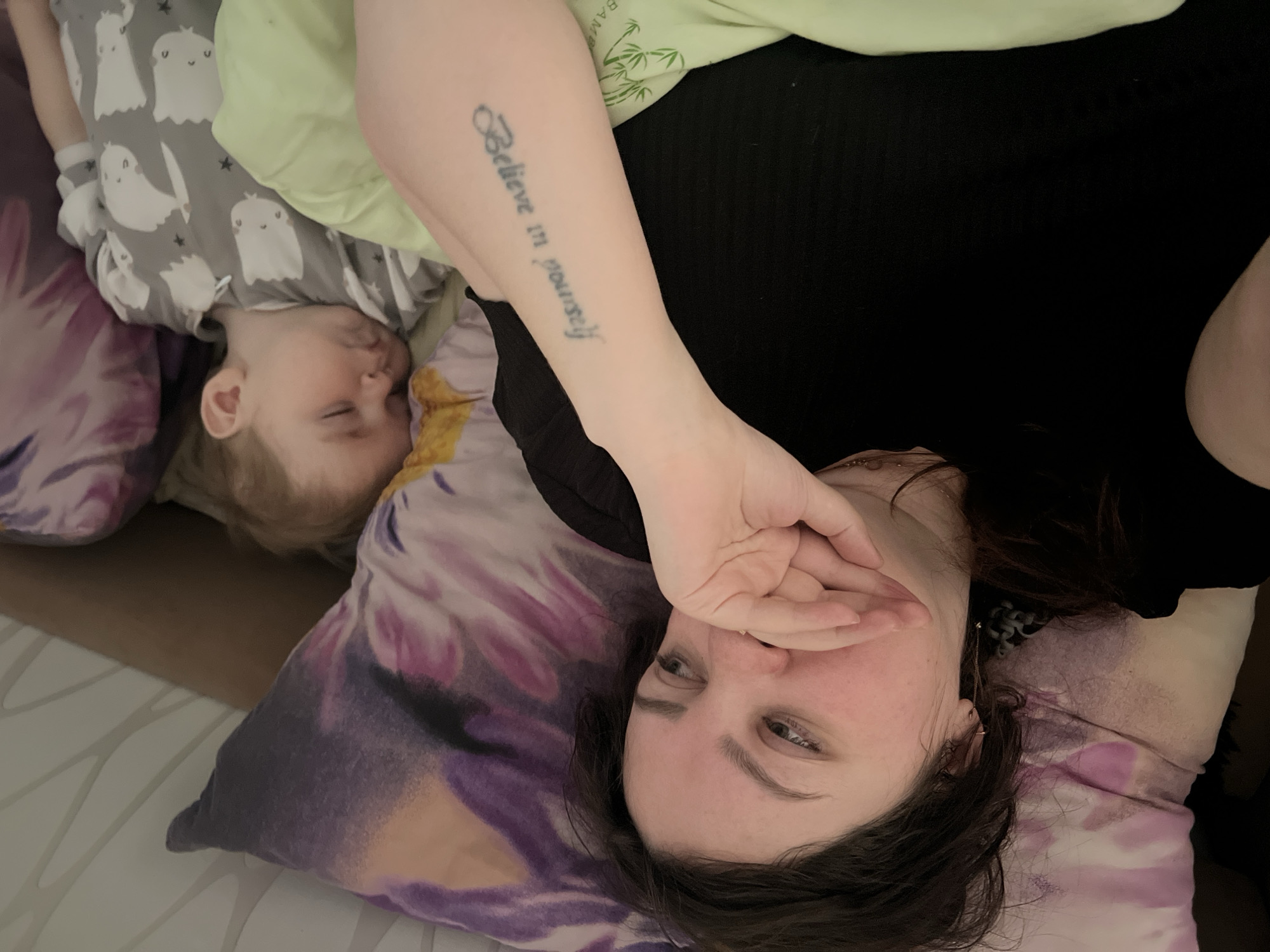 A woman and a young child are lying on pillows in an indoor setting. The child, wearing gray pajamas with a ghost pattern, is asleep. The woman, with brown hair, is partially covering her mouth with her hand and looking at the camera. Her left forearm is visible, displaying a tattoo that reads "Believe in yourself." The background includes a light green pillow, a purple patterned pillow, and what appears to be a wooden surface. I cannot confirm the location within the city of Perm, Russia.