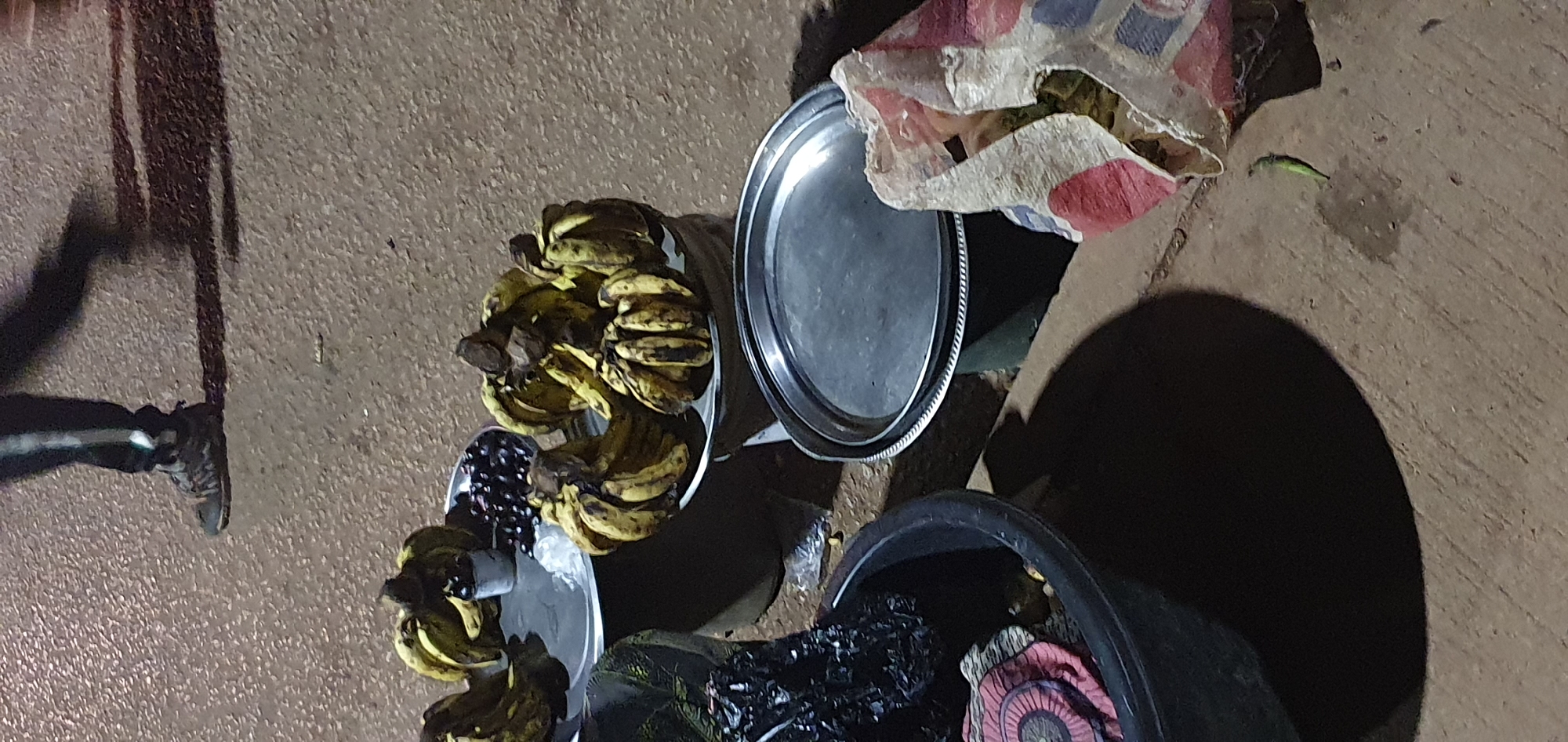 In Bukuru, Nigeria, a nighttime scene captures a street vendor's stall. The foreground is dominated by a large black basin containing a dark, crinkled material, possibly garbage or packaging, with a circular patterned cloth peeking out. To the right, a crumpled, colorful patterned bag lies open, revealing its contents. A single green chili pepper is visible on the ground.
Behind the basin and bag, three metal platters are arranged. The first platter on the left holds a bunch of bananas and a small cup filled with dark berries, possibly grapes or plums, next to a white substance, likely sugar or salt. The second platter, slightly behind and to the right, is piled with several bunches of overripe, yellow bananas, some showing brown spots. The third platter, in the background, also displays bananas.
In the upper portion of the image, a person's lower legs and feet are visible, suggesting they are walking away from the stall. They are wearing dark pants with a white stripe and dark, sturdy shoes. The ground is a rough, textured surface, likely a paved street or sidewalk. The lighting suggests artificial illumination, casting long shadows from the objects and the person.