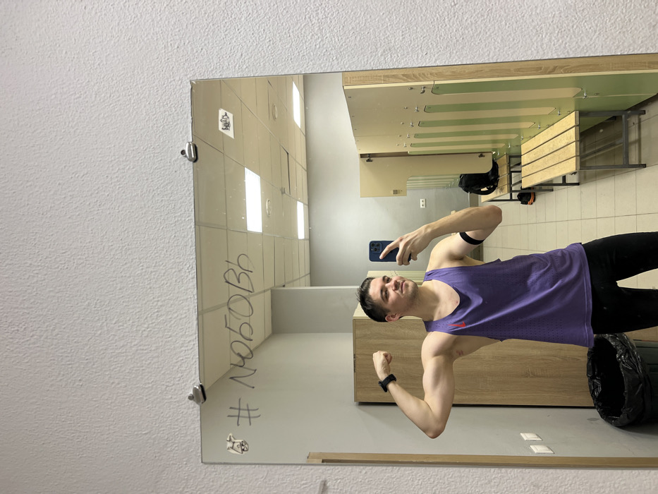 A man is taking a mirror selfie in what appears to be a locker room. He's wearing a purple tank top and black athletic pants, flexing his arm for the camera. The mirror is mounted on a wall with the hashtag "#любовь" written on it. The reflection shows lockers, benches, and other changing room elements. He is holding an iPhone in his left hand.