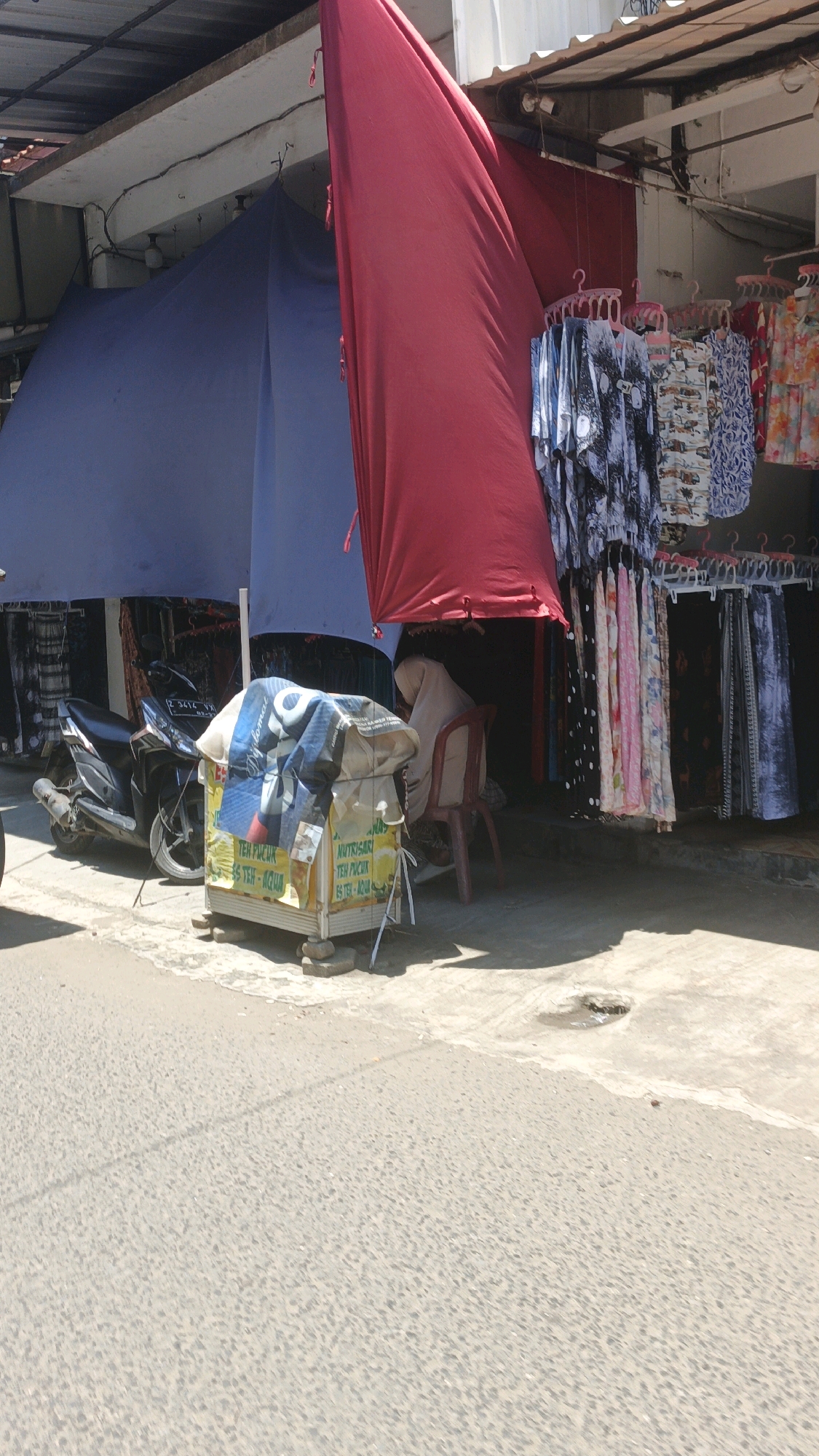 The image depicts an outdoor marketplace scene, likely in a rural or semi-urban setting given the open-air structures. A large red tarp is prominently displayed, partially covering a blue tarp that shelters a small motorcycle and what appears to be a vendor's stall. The stall itself is constructed from wood and is covered with various fabrics and signage, including text that reads "TEH PUCUK" and "NUTRISARI TEH". A person wearing a headscarf is seated behind the stall. In the background, clothing items are hanging on display, suggesting a retail area. The ground is paved with asphalt. The overall impression is one of a functional, albeit somewhat cluttered, commercial space operating under a bright, sunny sky.
