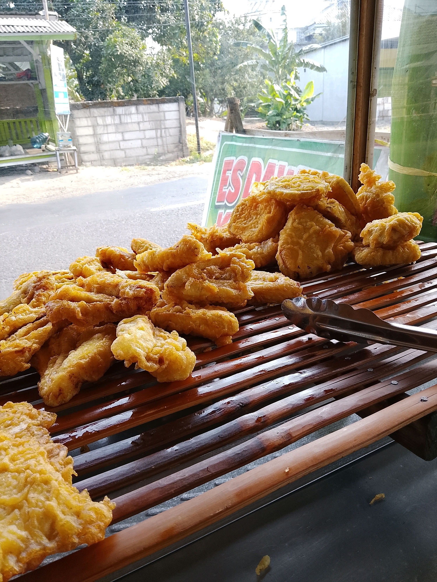 A close-up, eye-level, medium shot captures a pile of golden-brown, fried pastries, possibly bananas or sweet potatoes, resting on a slatted wooden surface. The pastries are irregularly shaped and appear to be coated in a crispy batter. A pair of metal tongs is positioned to the right of the pastries, suggesting they are being served or prepared. The wooden surface is slightly wet, with streaks of moisture visible. In the background, through a window, a street scene is visible, with trees, a brick wall, and a building. A green and white sign with Indonesian text is partially visible on the left. The lighting suggests an outdoor setting, likely during the day. The overall impression is of a street food vendor's stall in Malangsari, Indonesia.