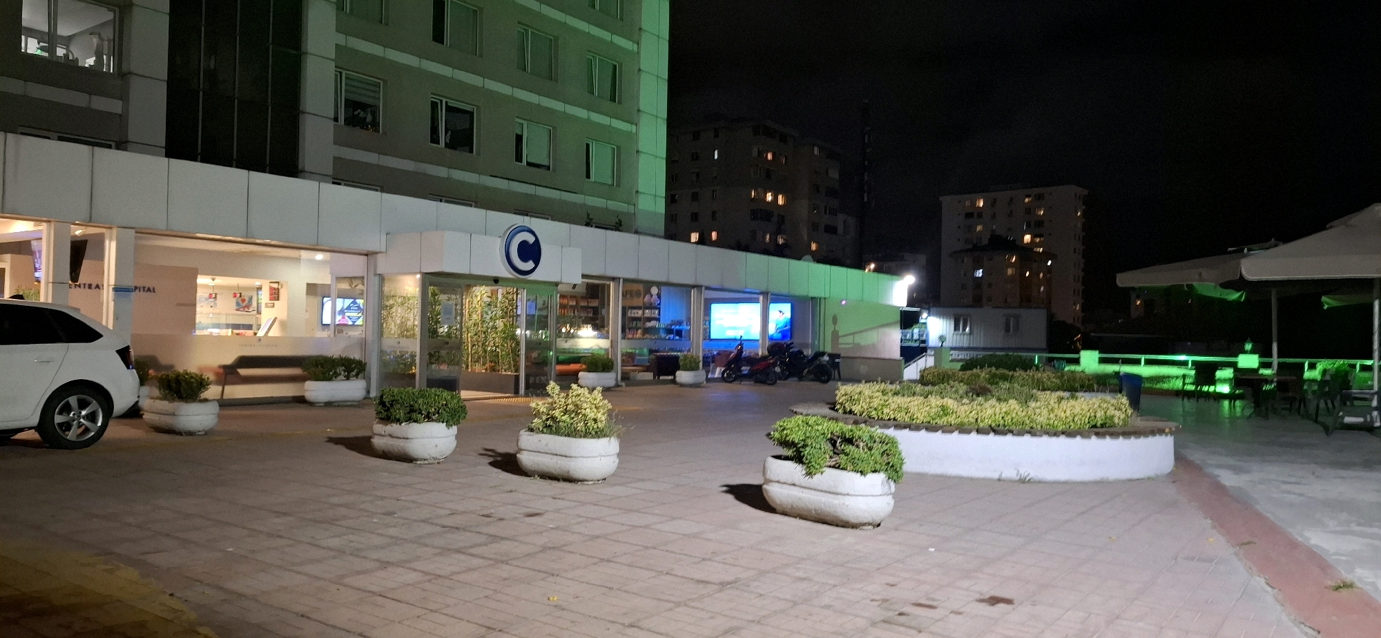 A nighttime exterior view of the Central Hospital in Kadıköy, Turkey, shows the building entrance, a white car, and surrounding outdoor seating areas. The hospital's facade features large windows and a prominent "C" logo above the entrance. Several potted plants are strategically placed around the entrance and in decorative planters. In the foreground, a white car is parked to the left. To the right of the entrance, a row of potted plants borders a seating area with tables and umbrellas, illuminated by green lights. Several motorcycles are parked near the entrance, and in the background, apartment buildings with illuminated windows are visible against the dark night sky. The ground is paved with interlocking bricks.