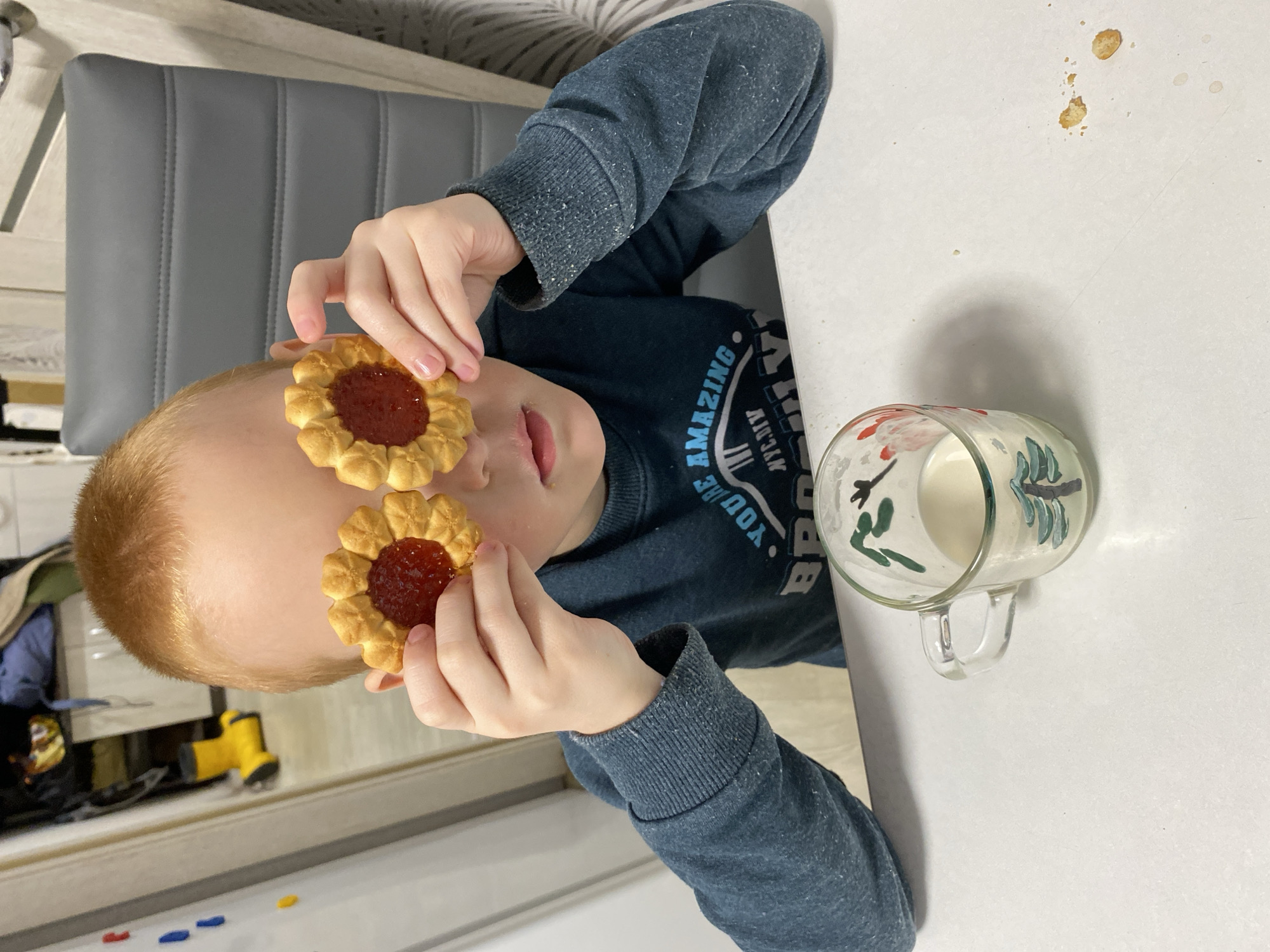 The scene depicts a domestic interior, identified as being in Noviy, Russia. A young, fair-skinned child with short, reddish-blonde hair is seated at a white table, centrally positioned in the frame. The child wears a dark blue long-sleeved shirt displaying a partial white graphic, including the text "YOU'RE AMAZING NYC." Both hands are raised to eye level, each holding a single flower-shaped cookie with a visible red jam filling, obscuring the child's eyes. The child's nose and mouth are discernible beneath the cookies. On the white table surface to the child's right, a clear glass mug with a green-painted tree design contains a small amount of white liquid, consistent with milk. Nearby, several small cookie crumbs and a faint brown liquid stain are present on the tabletop. The background on the right features a grey upholstered chair with horizontal stitching. The background on the left reveals a light-colored surface with small, colorful magnets, and on the floor below, a pair of yellow boots is partially visible.
