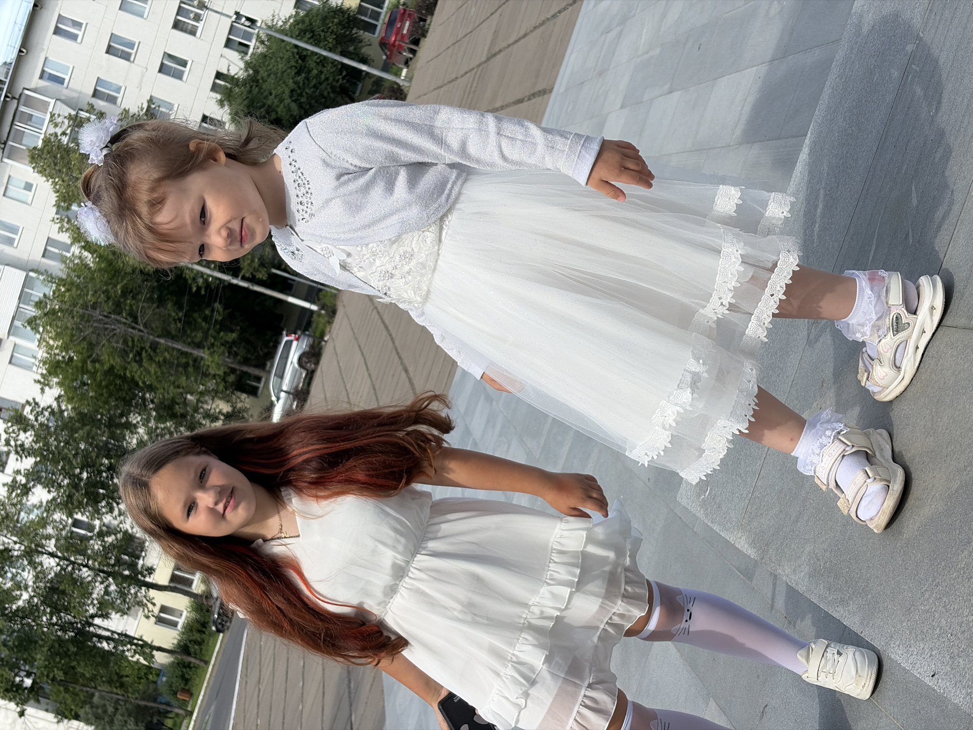 Two young girls, one older with long auburn hair and the other younger with blonde hair adorned with white hair bows, stand on concrete steps outdoors. The older girl wears a white dress and white socks with black cat designs, and the younger girl wears a white sparkly dress with a silver cardigan and white socks with lace trim. A multi-story building with white and yellow facades and numerous windows is visible in the background, along with trees and parked cars. The scene appears to be in an urban environment, likely a residential area.