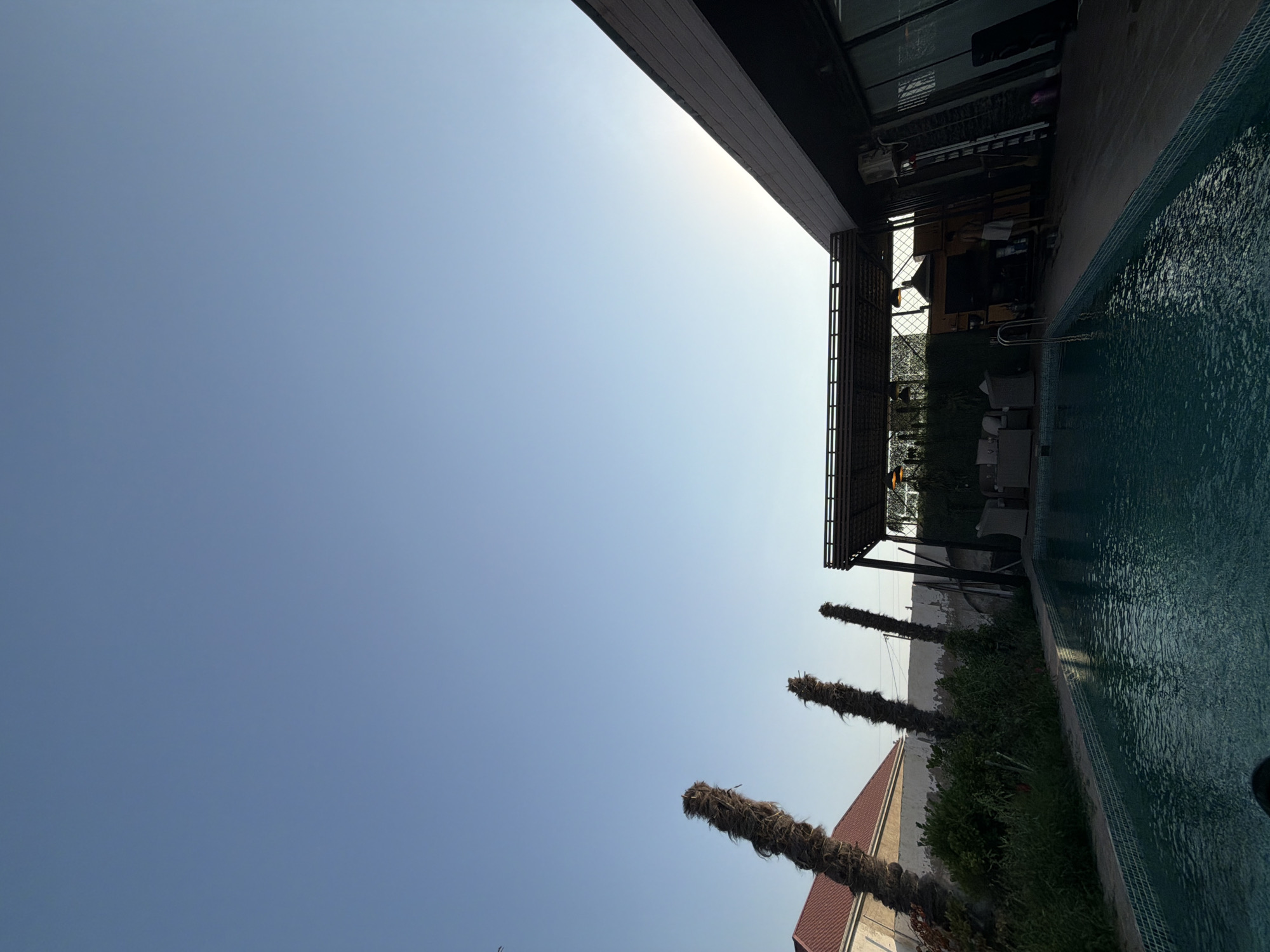 A photograph taken in the city of Ləhiş bağları, Azerbaijan, shows an outdoor swimming pool area with a modern building and some palm trees against a clear blue sky. The pool is filled with turquoise water and has a tiled edge. To the left of the pool, there are three dead palm trees, one of which is significantly taller than the others. Beyond the palm trees, a building with a red tiled roof is partially visible. To the right of the pool, a dark, contemporary building with large windows and a sloped roof is present. An outdoor seating area with wicker furniture is situated under a covered patio structure. A person, appearing to be a male wearing shorts, is visible standing on the deck of the modern building, near a ladder and some cleaning supplies. The overall impression is of a resort or private residence in an arid climate.