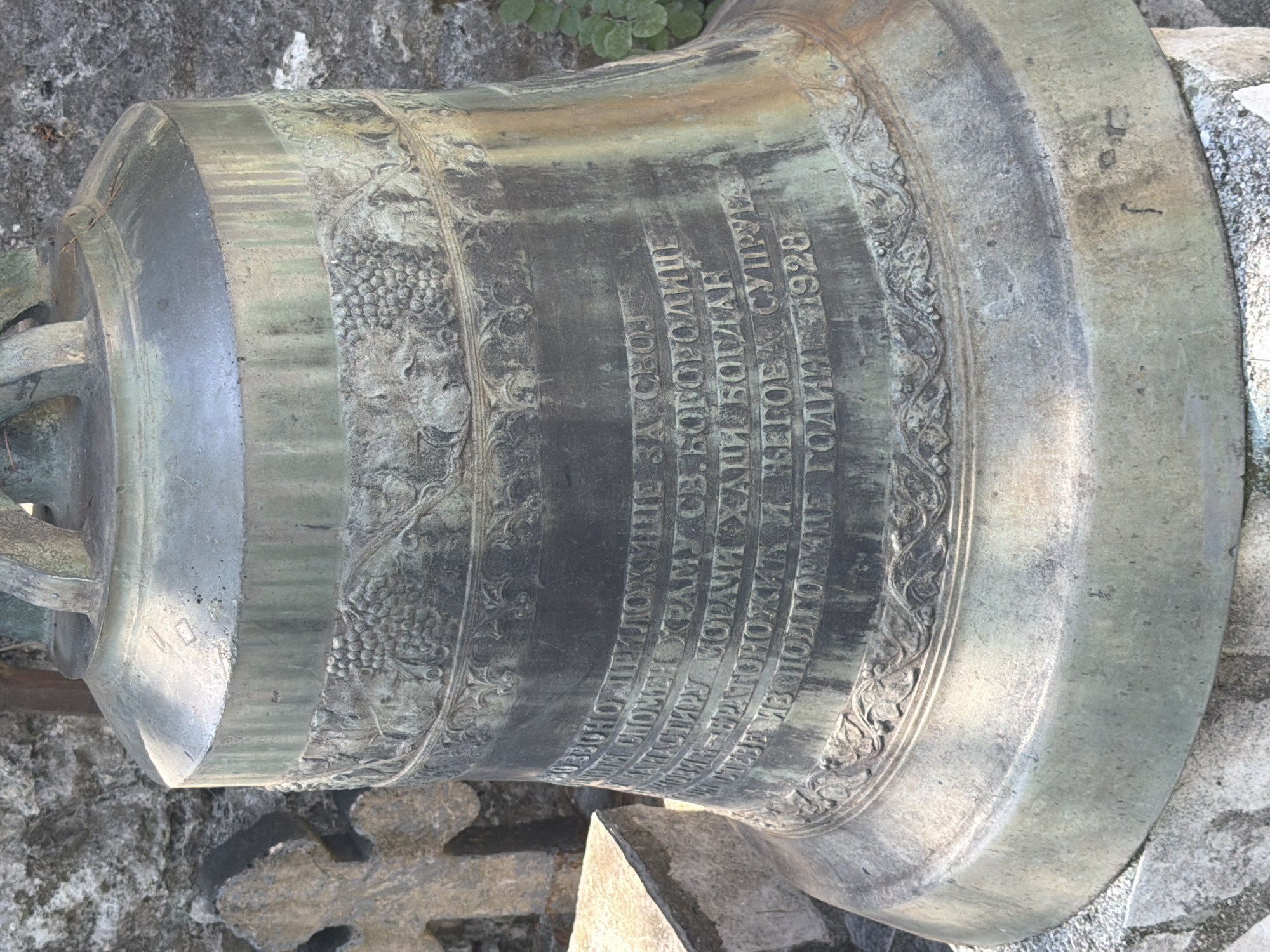 The image shows a close-up view of a large, aged bronze bell. The bell is ornately decorated with relief carvings of grapes and floral patterns. Prominently displayed on the bell's surface is an inscription in Cyrillic script. The inscription appears to be a dedication or memorial. The bell is situated outdoors, with a rough stone wall and a stone cross visible in the background. Some green foliage is also present on the right side of the frame. The overall impression is one of historical significance and weathered permanence. The inscription refers to the city of Manastir Morača, Montenegro.
