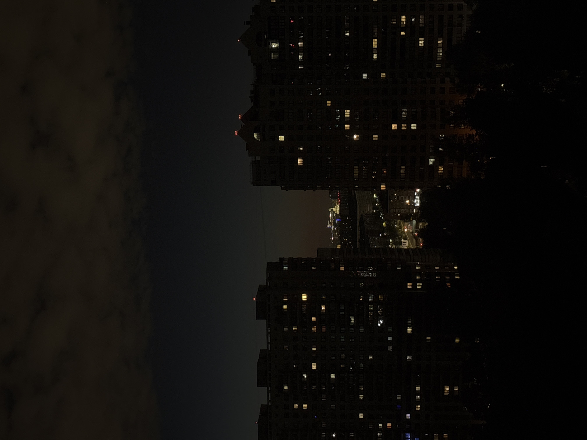 This is a nighttime, high-angle shot of a city skyline. Two large apartment buildings dominate the foreground, silhouetted against a dark sky. Numerous windows in the buildings are lit, indicating occupancy. The building on the left is taller and appears to have a flat roof with some lights, while the building on the right has a more complex roofline with red lights on two peaks.
In the background, a sprawling city with more lights is visible, suggesting a densely populated area. Some trees are visible in the dark foreground, obscuring the lower portions of the buildings. The sky is dark with some cloudy formations illuminated by ambient light. The overall impression is one of a quiet, urban night.