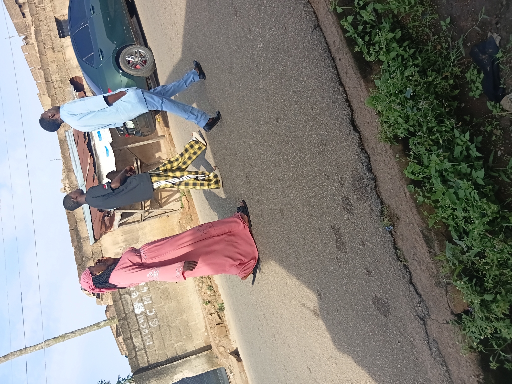 Three people are walking on a street in front of a brick wall. A woman in a pink dress and headscarf walks on the left, followed by a man in a black t-shirt and yellow plaid pants. To the right, a man in a light blue shirt and pants walks. A dark green sedan is parked behind the people, partially visible. The street is paved with asphalt and there is a curb with green foliage. The sky is visible in the background, with a utility pole on the left.