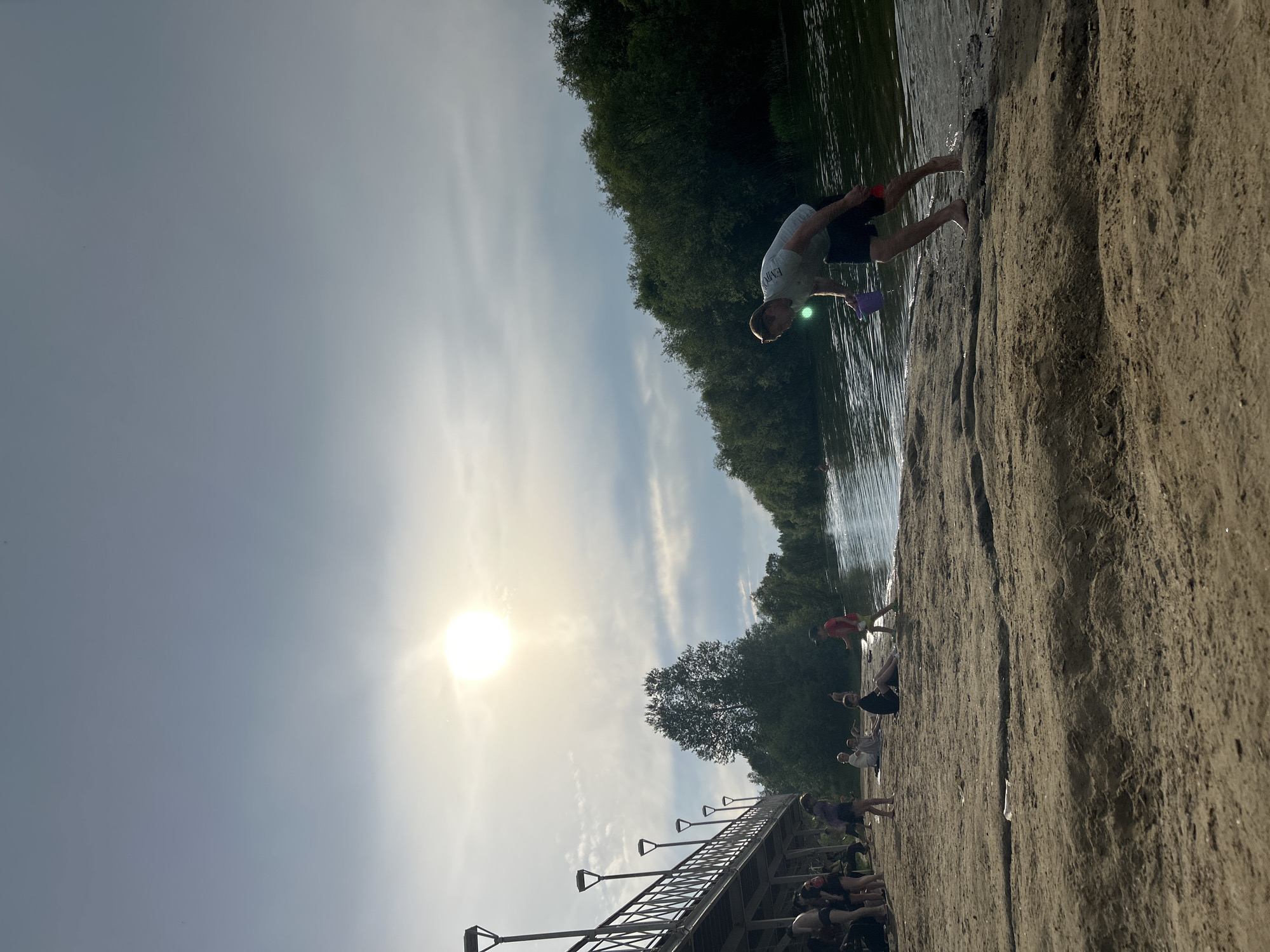 The image depicts a sunny day at a sandy beach next to a body of water, likely a river, as suggested by the presence of trees lining the opposite bank. In the foreground, a young man is bending down at the water's edge, holding a purple bucket and a red object, possibly a shovel. His legs are submerged in the water, indicating he is either entering or exiting the water. In the midground, several people are scattered across the beach. Some are sitting, while others appear to be standing. On the left side of the image, a pedestrian bridge with lampposts runs parallel to the shoreline. The sky is partly cloudy with the sun shining brightly, creating a hazy effect. The overall scene suggests a recreational setting, possibly a public beach. The specific location is identified as Angarsk, Russia, and the scene may be the beach area near the Angara River, a common recreational spot in the city.