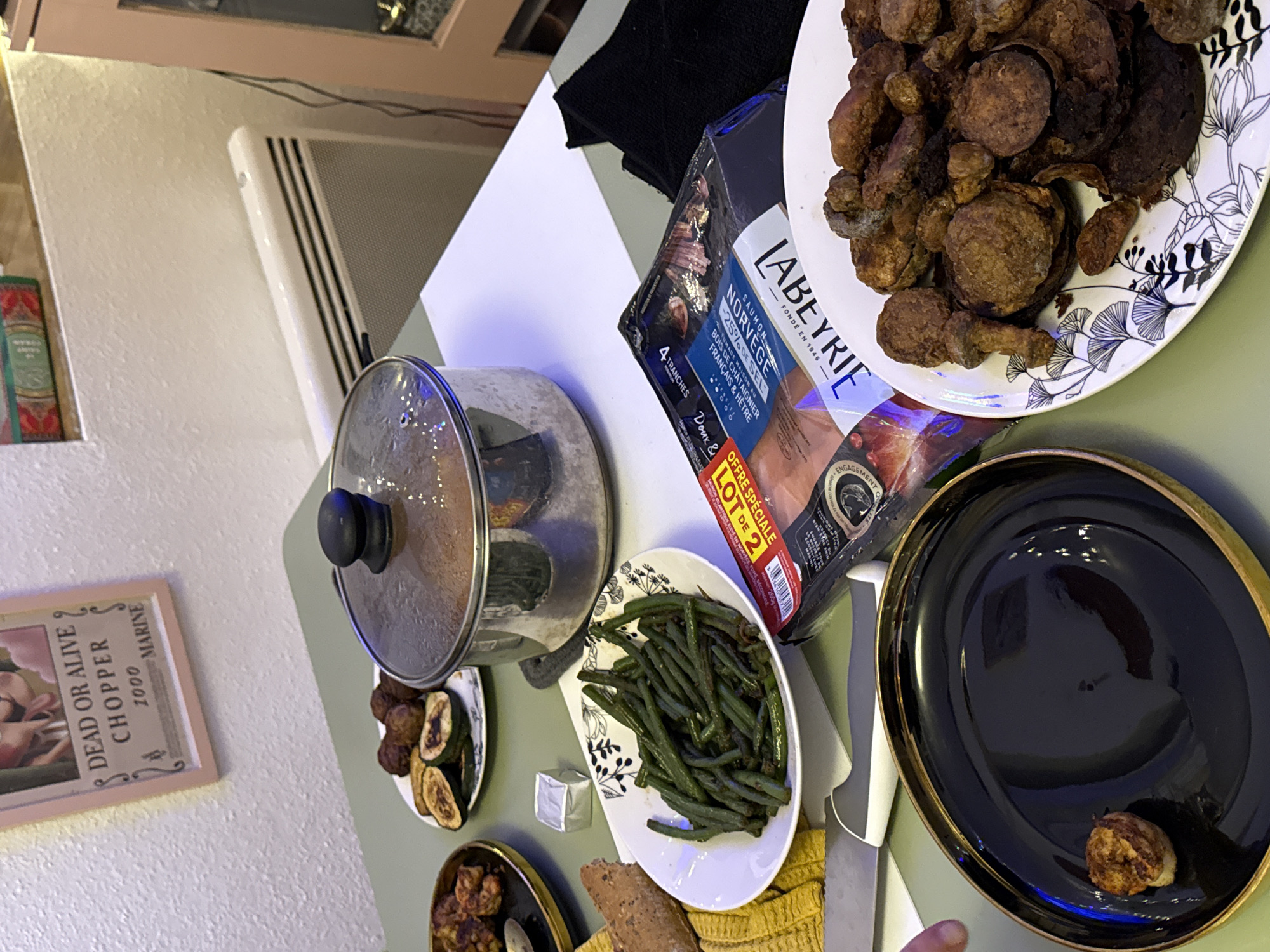 This image captures an indoor scene, likely a dining area, within the city of Strasbourg, France.
A light green tabletop serves as the primary surface, laden with a variety of prepared food items and associated objects. In the center, a stainless steel pot with a transparent glass lid, showing condensation, suggests a hot dish. To its left, a white oval plate contains a serving of dark green beans. Adjacent to the green beans is a small, foil-wrapped square, possibly butter or a bouillon cube. Further left, a piece of crusty bread rests on a yellow, textured towel. A kitchen knife with a white and grey handle is positioned below the towel.
In the bottom-left quadrant, a black, glossy plate with a gold rim holds a single, irregularly shaped, browned food item. A human hand, showing two fingers, is partially visible near this plate. To the right of the pot, a vacuum-sealed package of "LABEYRIE Saumon Fumé de Norvège" (Smoked Salmon from Norway) with "4 tranches" (4 slices) and "OFFRE SPECIALE LOT DE 2" (special offer lot of 2) is placed on a white surface, possibly a placemat, next to a folded black cloth. Below the smoked salmon package, a white plate with a dark leaf pattern is filled with numerous dark brown, irregularly shaped, cooked items resembling fried mushrooms or croquettes. Another white plate further back to the left holds several round meatballs and slices of roasted zucchini.
The background consists of a textured, off-white wall. On the wall, a light pink-framed picture displays a "WANTED" poster-style image for "DEAD OR ALIVE CHOPPER" with a bounty of "1000 MARINE," referencing the character Tony Tony Chopper from One Piece. To the right, a white wall-mounted electric heater or radiator is visible, with a small, red-bound book or object resting on a narrow shelf above it. A portion of a light-colored cabinet with a glass door is also visible on the far right. The scene is illuminated by ambient indoor lighting.