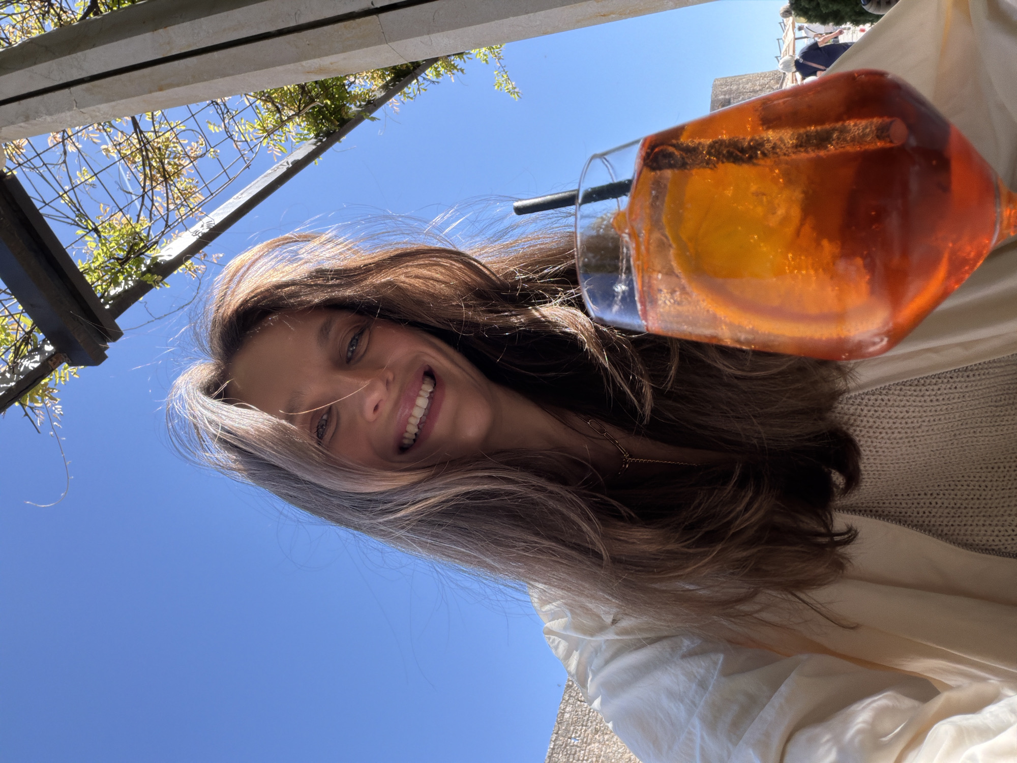 A person with long brown hair is smiling and looking directly at the camera while holding a tall, angular glass filled with an orange-colored beverage and a black straw. The beverage appears to contain a slice of orange. The background includes a clear blue sky, a pergola-like structure with greenery, and a building in the distance. The person is wearing a light-colored top. Given the presence of the pergola, the outdoor setting, and the type of drink, the photo may be taken at a cafe or restaurant in Budva, Montenegro, though this is only speculative and cannot be definitively confirmed from this single image.