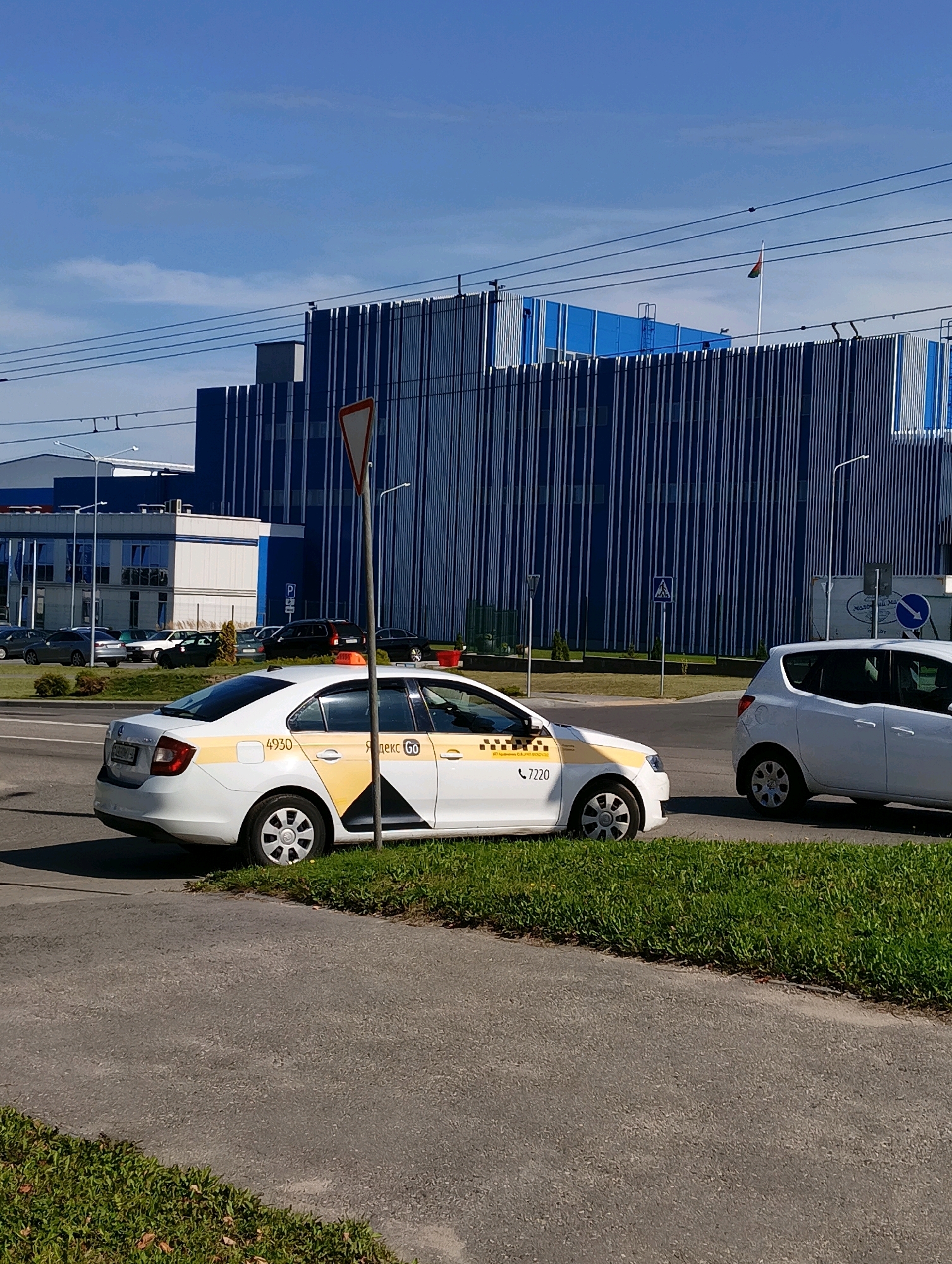 A white taxi with "Yandex Go" branding and the number 4930 is parked on the grass verge next to a paved road. The taxi is a Skoda Rapid and has a yellow stripe on its side. Another white car, possibly a hatchback, is parked further down the road. In the background, a large blue building with vertical white lines dominates the skyline, with a Belarusian flag flying on a pole. The sky is clear and blue. The scene appears to be in an industrial or commercial area.