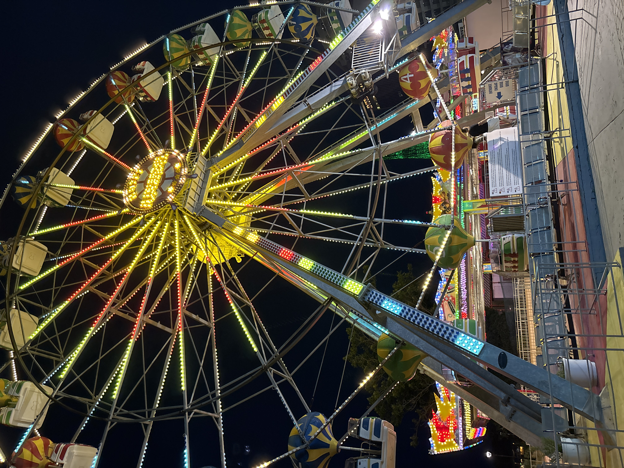 A close-up, low-angle shot captures the illuminated Ferris wheel at the Varna Sea Garden amusement park at night. The wheel is adorned with a multitude of multicolored LED lights, primarily yellow and white, with accents of red and green. Several gondolas, designed to resemble hot air balloons with yellow, red, and blue patterns, are attached to the wheel's spokes. The central hub of the Ferris wheel features an illuminated sign. In the foreground, rows of empty metal seats and safety railings are visible, suggesting a break in operation or the early stages of the park's opening. The background is a dark night sky, with some trees and other amusement park rides subtly visible.