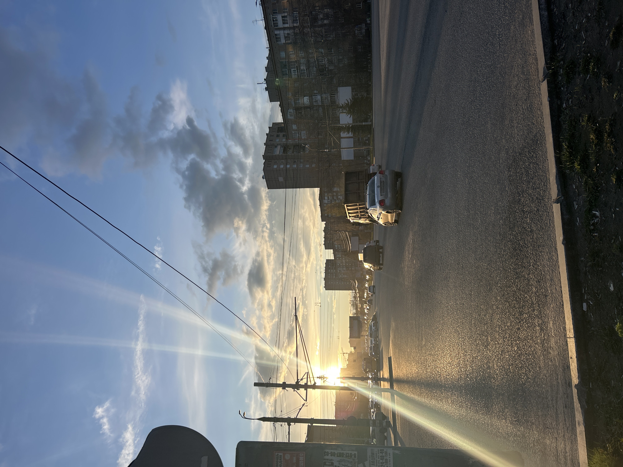 The image captures an urban street scene at either sunrise or sunset, characterized by warm, golden light and long shadows. The camera is positioned low, looking along an asphalt road that appears vertically oriented due to the original image's portrait capture and subsequent rotation. The bright sun, low on the horizon, is a dominant feature, casting intense rays and lens flares across the scene and reflecting strongly off the wet-looking road surface.
The sky above is partly cloudy, displaying patches of clear blue interspersed with white and grey clouds. Several utility poles with a network of overhead wires crisscross the frame, particularly prominent in the foreground and mid-ground, suggesting infrastructure for public transport or power lines. Multi-story apartment buildings, appearing somewhat aged, line the left side of the street, partially silhouetted against the bright sky and obscured by the sun's glare.
Several cars are visible on the road, including a silver sedan (partially obscured) in the mid-ground, and others further in the distance appearing as bright reflections. A grassy strip with some sparse vegetation runs alongside the road, separated by a concrete curb. In the bottom right foreground, a dark green utility box or sign structure is visible, featuring Cyrillic text. Readable elements include "Реклама" (Advertisement), "АВТО" (Car), "УЛ. ЛЕНИНА" (Lenin Street), and various phone numbers such as "8-912-607-00-03" and "24-40-00", reinforcing the Izhevsk, Russia, location context. The top of a person's head or a car's dashboard is partially visible in the bottom left corner, indicating the photographer's perspective. The scene conveys a tranquil, everyday moment in an urban setting bathed in the dramatic light of the rising or setting sun.