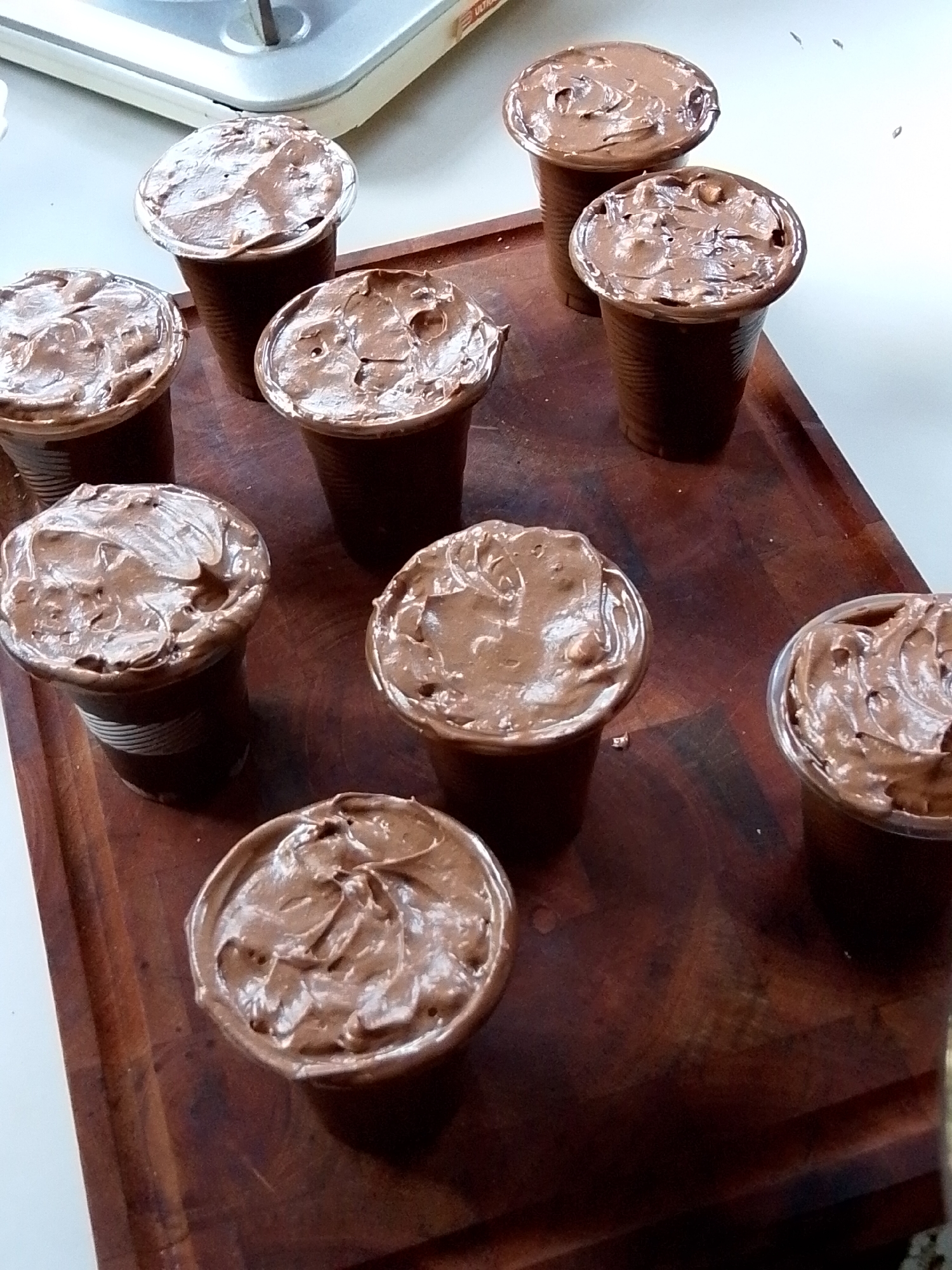 The image displays a close-up overhead view of a dark wooden cutting board with several small, clear plastic cups filled with a rich, brown, mousse-like dessert, possibly chocolate mousse or pudding. The dessert in each cup is swirled with a spoon, creating peaks and valleys. The cups are arranged in a staggered pattern on the cutting board, with some closer to the foreground and others further back. The lighting suggests an indoor setting, possibly a kitchen. The background is out of focus, showing a white surface and a metallic object with a handle, which could be a kitchen appliance. There is no visible indication of the city of Davao City, Philippines, within the image itself, nor are there any people or specific actions or interactions depicted.