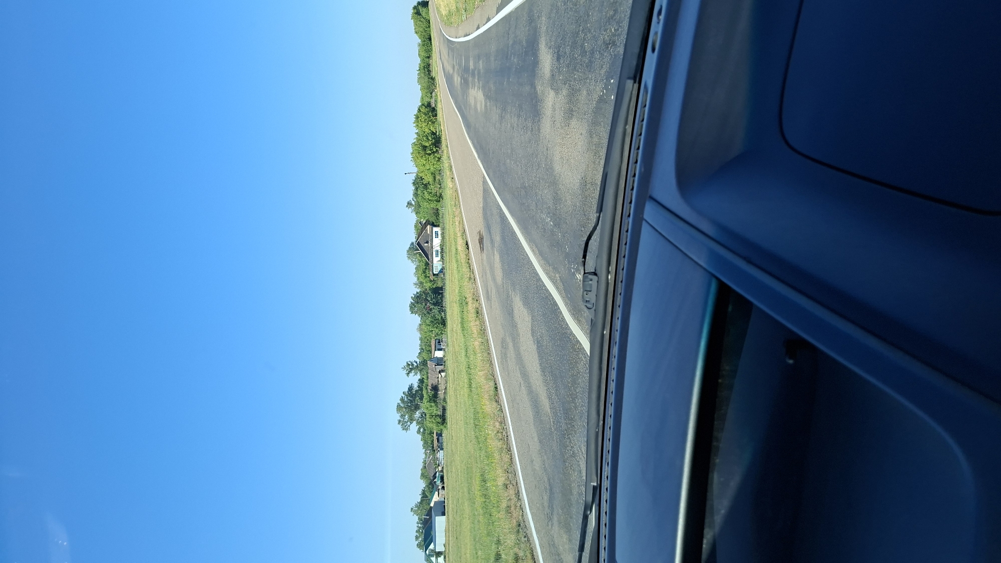 The image depicts a view from inside a vehicle, looking out through the windshield at a rural road. The road is paved and has white lines indicating lanes. On either side of the road are green fields with scattered houses and trees. The sky is clear and blue. The foreground of the image shows the dashboard of the vehicle. There are no people or animals visible in the image. The scene appears to be a sunny day in a rural area.
