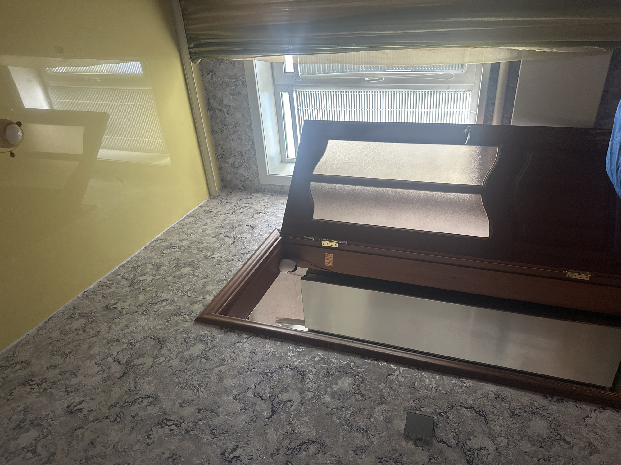 The image depicts an interior view of a room, possibly an apartment, in Leninsk-Kuznetsky, Russia. The room features textured, grey and dark blue patterned wallpaper on one wall. The ceiling is a smooth, reflective yellow material, showing reflections of a window with blinds and a light fixture. A dark wooden door is ajar, revealing a tall, narrow mirror or a reflective surface inside an adjoining space. The door has frosted glass panels with a wooden frame and a metal handle. A small white security camera is visible on the wall near the door frame. Next to the door on the wall is a small black electronic device. Sunlight streams in through a white-framed window covered with horizontal blinds, and a light-colored curtain hangs on the right side of the window. A radiator is visible below the window. A portion of a blue blanket or fabric is in the lower foreground.