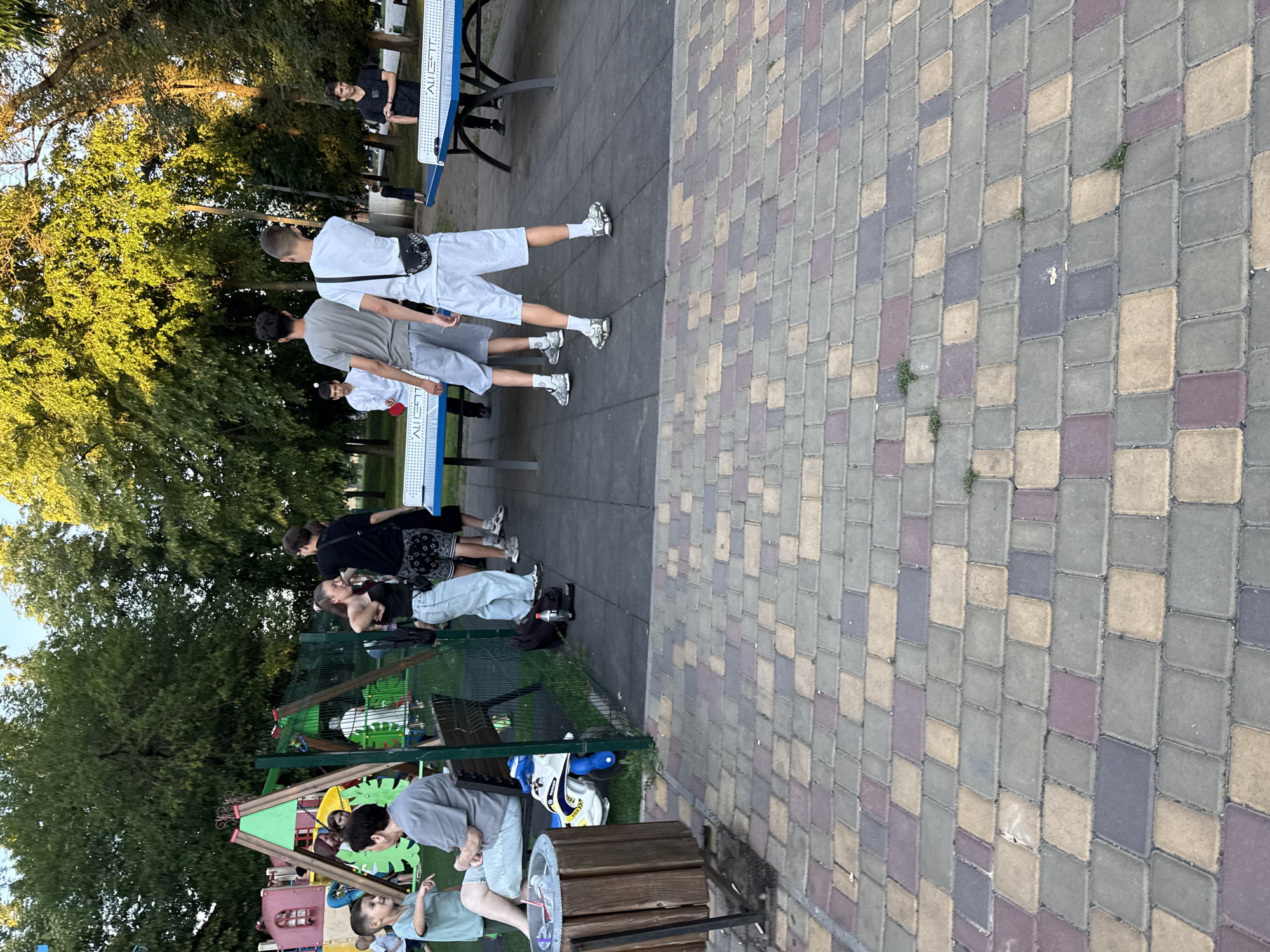 In an outdoor recreational area, several young people are gathered. On the left, a child sits on a bench near a playground structure. In the center and right, groups of young men and women are engaged in playing ping pong. Two ping pong tables are set up on a dark grey paved surface. Trees with green foliage surround the area, suggesting a park or public space. The ground is paved with colorful rectangular bricks.