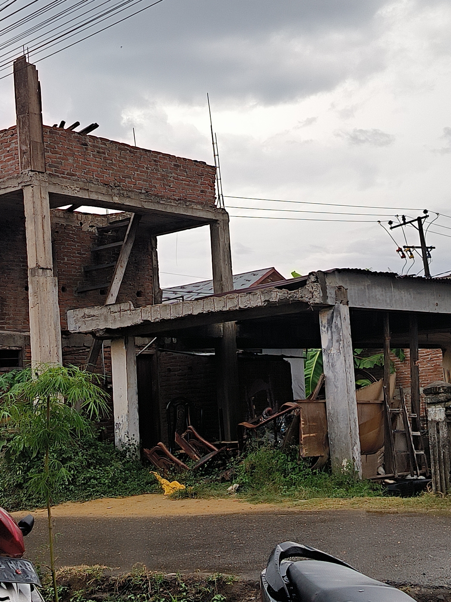 The image captures an unfinished or dilapidated two-story building made of concrete and red bricks, set beside a paved road under an overcast sky. The building's concrete frame, featuring exposed columns and beams, is prominent, with segments of red brick wall visible on both levels. Rebar extends upwards from the concrete structures, indicating incomplete construction. A simple wooden staircase can be seen within the upper floor.
The ground floor area beneath the structure is cluttered with various discarded items, including broken wooden chairs, wooden panels, and other debris, amidst dense green vegetation and weeds. A small bamboo plant stands in the left foreground near the roadside.
In the immediate foreground, parts of two motorbikes are visible: the front of a red motorbike with a partially obscured license plate ("DL XXX X" is discernable) on the left, and the black seat and rear of another motorbike on the right. Overhead, power lines span the cloudy sky, connected to a utility pole on the right side of the frame. The absence of direct sunlight suggests an overcast day. No people are present in the scene, which conveys a sense of abandonment or a prolonged halt in construction, typical of an urban or semi-urban setting in Lhokseumawe, Indonesia.