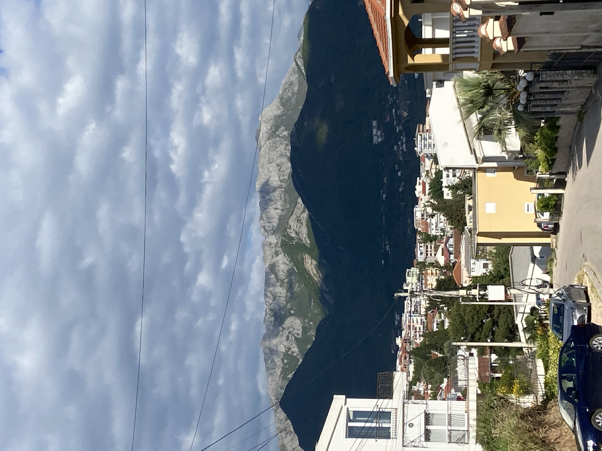 This image, when correctly oriented, presents a scenic view of a coastal town nestled against a dramatic mountainous backdrop under a partly cloudy sky.
In the background, a large and imposing mountain range dominates the scene. Its upper reaches feature light-colored, rugged rocky peaks, while the lower slopes are covered in dense, dark green forest. The midground is filled with numerous white and light-colored buildings, characteristic of Mediterranean architecture, many with red or orange tiled roofs. These structures appear to be terraced, climbing up the hillside towards the mountains. Green foliage, including several prominent palm trees, is interspersed among the buildings, adding a lush contrast.
In the foreground and lower right, residential buildings with ornate balconies and windows are visible. A paved road curves through this area, where several cars are parked, including a dark blue sedan. Utility lines or power cables stretch across the sky in the upper portion of the frame.
The weather appears to be daytime, with a mix of blue sky and soft, fluffy white clouds. The overall atmosphere is bright and serene, suggesting a pleasant day. The architectural style and mountainous coastal setting are consistent with the location context of Šušanj, Montenegro. There is no visible text in the image.