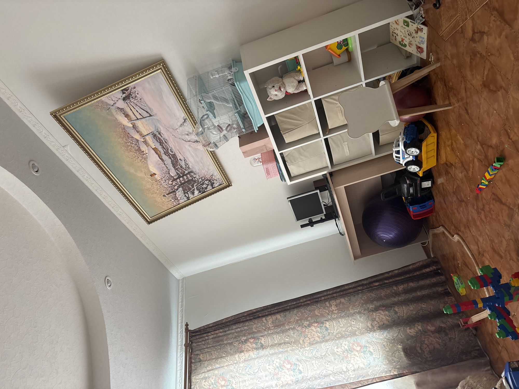 The image shows an indoor scene, possibly a child's room, with various objects and furniture. On the left, a framed painting with a gold-colored frame hangs on the wall. Next to it is a birdcage. Adjacent to the birdcage is a white shelf unit with storage bins and toys inside, including a stuffed cat. Beside the shelf unit, there's a desk with a computer monitor and a purple exercise ball underneath. On the floor, there are several toy trucks, colorful building blocks, and other toys scattered around. In the background, a window with a patterned curtain is visible. The specific location in the city of Ferma, Russia, cannot be determined from the image.