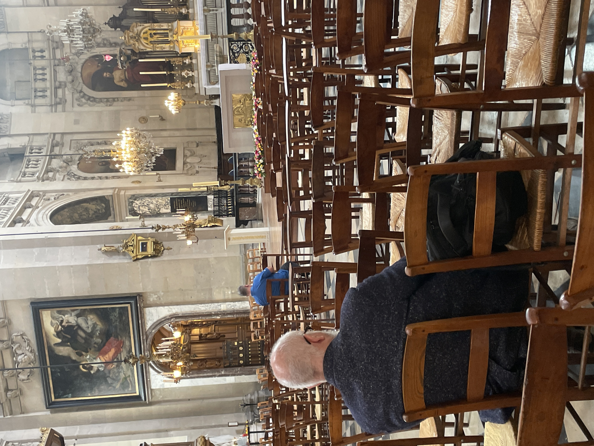 The image depicts the interior of a grand church, likely in Paris, France, given the architectural style and ornate decorations. In the foreground, an older man with a bald head and glasses is seated, facing away from the camera, towards the altar. He is wearing a dark, textured jacket. Behind him, rows of empty wooden chairs with woven seats are arranged. Further back, another person in a blue shirt is seated in a chair, also facing the altar. The church features high ceilings, elaborate chandeliers, and religious artwork displayed on the walls. The altar area is decorated with flowers and a golden relief, with large candles and an organ visible in the background. The overall atmosphere is one of quiet reverence and solemnity, common in religious services.