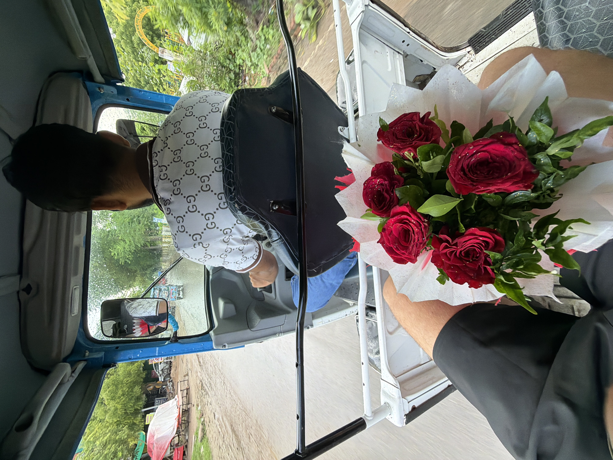 The image shows the interior of a blue autorickshaw in Kanpur, India. A man wearing a white t-shirt with a repeating "GG" pattern is driving. In the foreground, a bouquet of red roses wrapped in white paper is visible. The autorickshaw is on a wet, unpaved road, suggesting it may have recently rained. Lush green trees line the road, and in the background, there are market stalls with covered goods and a distant yellow archway. The reflection in the driver's side mirror shows another autorickshaw in the distance. The perspective is from inside the autorickshaw, looking forward and slightly to the right.