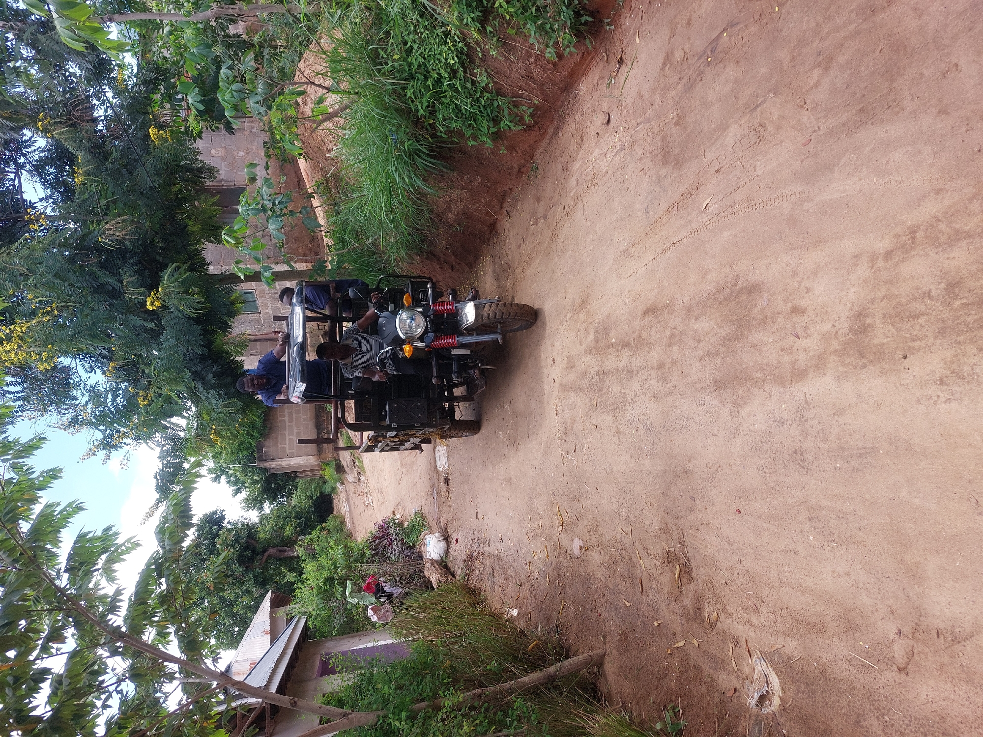 The image shows a red dirt road in what appears to be a rural area. A three-wheeled vehicle, likely a tuk-tuk, is on the road, carrying several passengers. Vegetation lines both sides of the road, including trees and tall grasses. A building is visible on the left side of the road, partially obscured by trees. The image appears to have been taken in a developing country, based on the dirt road and the presence of the tuk-tuk. It is not possible to confirm whether this is in Dar es-Salaam, Tanzania, based on the image alone. More specific details would be needed to confirm the location.