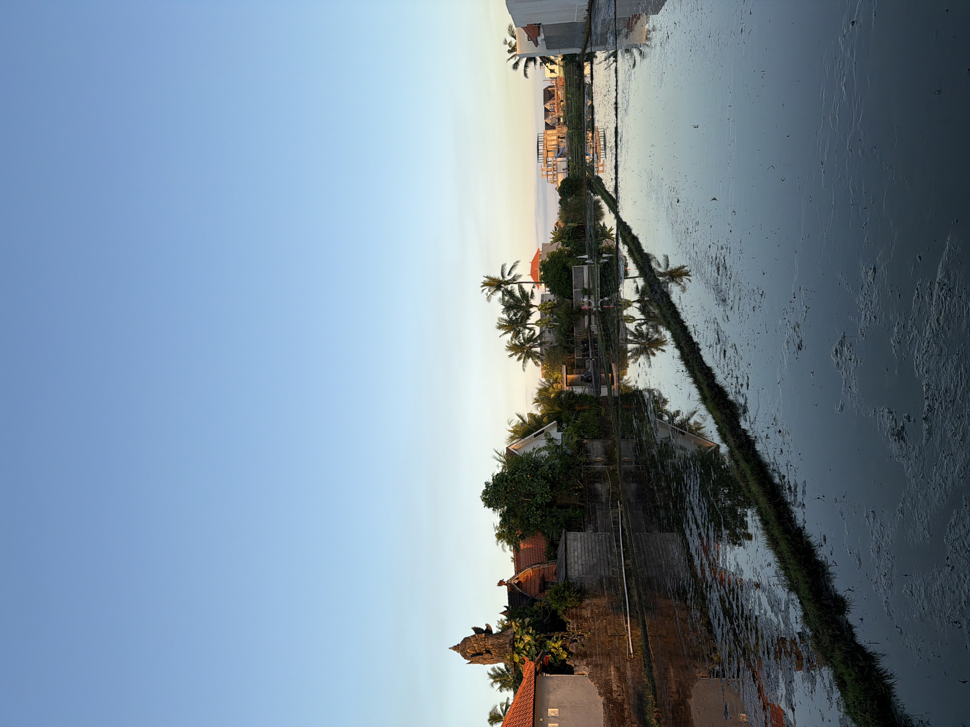 The image shows a landscape in Mengwi, Indonesia, reflected in a still body of water, likely a rice paddy. The composition is divided between a clear, light blue sky on the left and the mirrored scene on the right. Buildings with terracotta roofs and surrounding greenery, including palm trees, are visible along the horizon. A raised embankment covered with grass divides the reflective water from the buildings. Distant structures under construction can be seen further into the scene. The time of day appears to be dusk or dawn due to the soft lighting and pastel colors. The water surface shows slight surface disturbances, blurring the reflection in some areas.