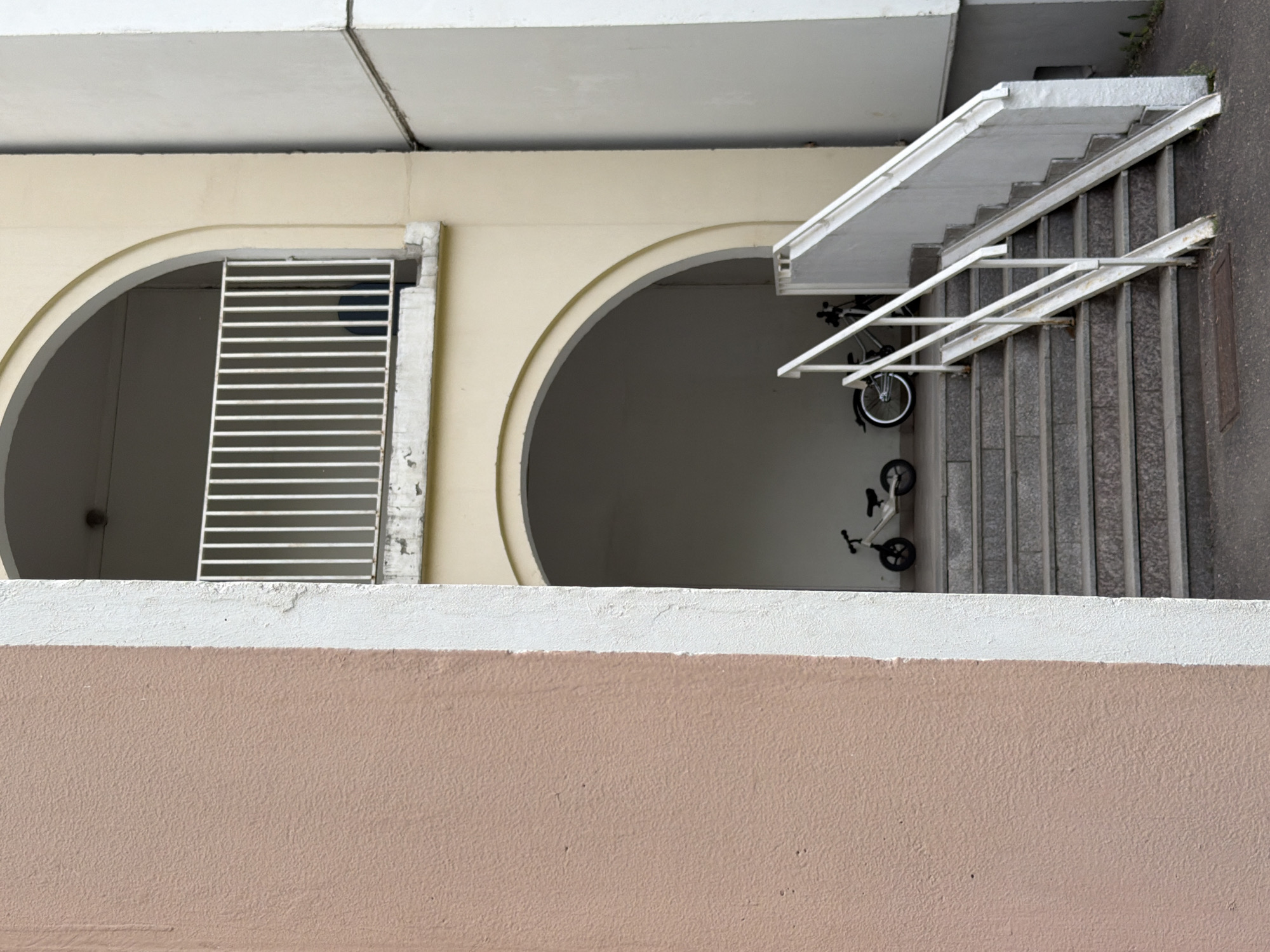 This elevated shot captures a functional architectural space in Khimki, Russia, featuring a neutral and utilitarian aesthetic. The composition looks down upon a section of a building or a covered exterior walkway.
In the immediate foreground, a textured, light brown (or peach) wall, possibly a parapet, spans the lower third of the image, topped by a crisp white coping. Above this, a light beige wall presents two prominent arched openings. The arch on the left is partially covered by a white horizontal slatted gate or barrier. Within the shaded area of the right arch, two small bicycles—one appearing to be a child's balance bike and the other a small two-wheeled bike—are parked on the ground.
To the right of these arches, a set of grey concrete stairs descends, equipped with a white metal handrail that extends towards the viewer. The ground adjacent to the stairs is a dark grey surface, likely asphalt, and a small, dark rectangular plate is embedded within it. Overhead, a white concrete ceiling or the underside of an upper level is visible, marked by panel lines. The lighting suggests it is daytime with soft, indirect illumination, as strong shadows are absent, possibly indicating an overcast sky or deep shade. There are no people visible, and no text is discernible within the frame. The scene conveys a sense of quiet urban utility.