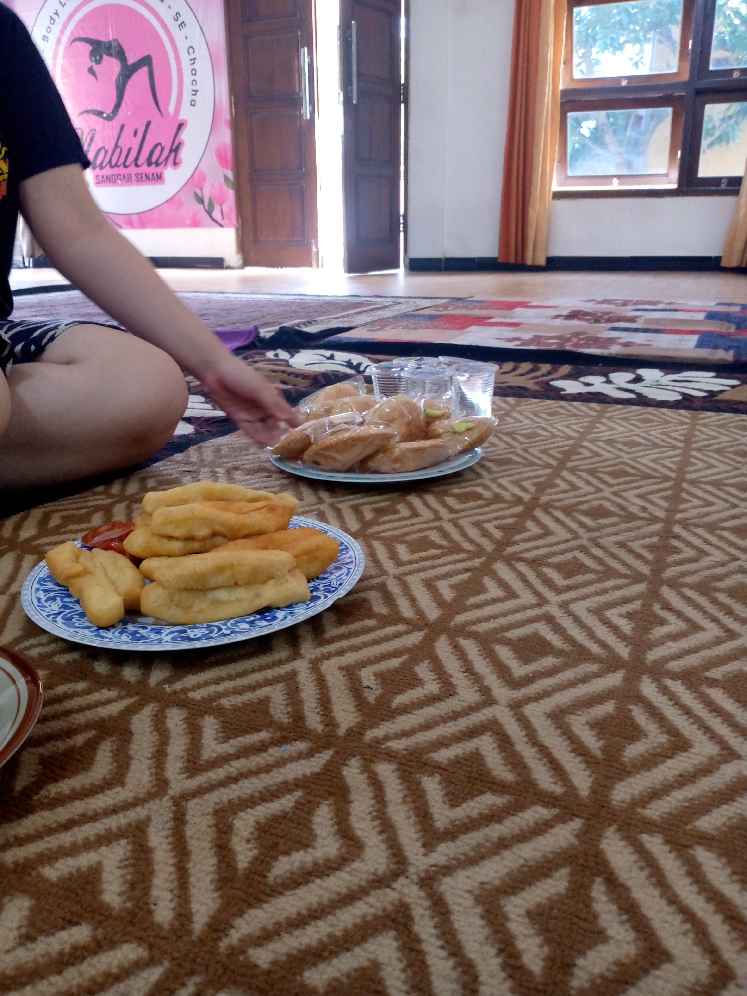 The image shows a person's leg and arm, along with two plates of food, on a patterned carpet. The food consists of fried dough pastries, one plate with a side of sauce. In the background, there are open doors and windows with curtains. A banner with text and an image is visible on the left. The scene appears to be indoors, possibly a recreational space or a home. The overall lighting suggests daytime. The banner on the left appears to be related to a dance studio or fitness center, with Indonesian text like "LABILAH" and "SANGGAR SENAM". Given the context of the banner, the location is likely an indoor space associated with a fitness or dance studio in Mojokerto, Indonesia.