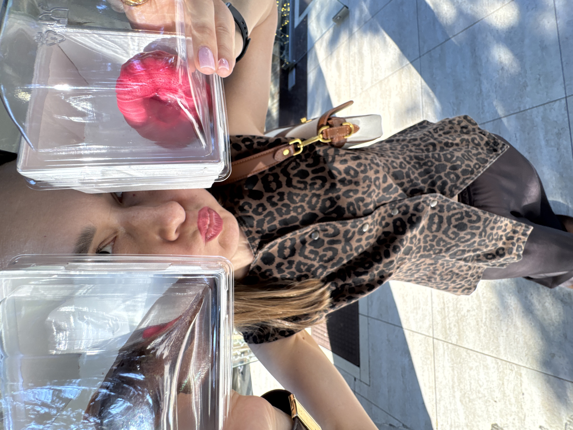A person is taking a selfie outdoors, holding two clear plastic containers, each containing a food item. The container on the left holds a dark, possibly chocolate, item, while the one on the right contains a bright red, textured item. The person is wearing a sleeveless top with a leopard print and dark pants, and a light-colored purse with a brown strap is slung over their shoulder. The background appears to be an outdoor tiled walkway with sunlight casting shadows. The overall impression is of a casual, sunny day. The provided information about the location being Sochi, Russia, is not visually verifiable from the image content itself.