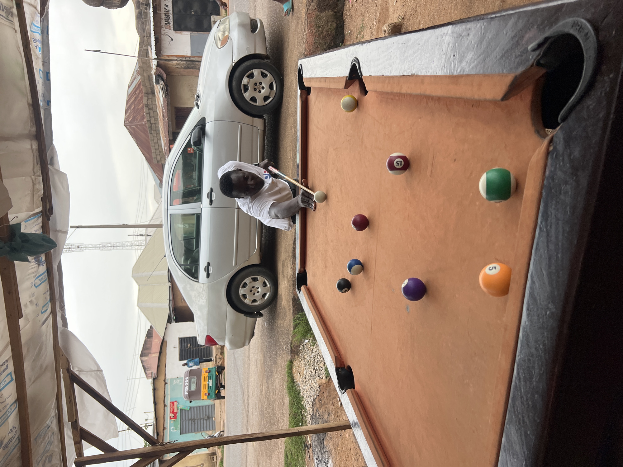 The image depicts a man playing pool beside a white car in Jos, Nigeria. The pool table is orange with black pockets and several pool balls are scattered across it. The man, who is Black, wears a white shirt and is focused on lining up his shot with a cue stick. A white car is parked next to the pool table on a paved area.
In the background, there are buildings of Jos, Nigeria. One has a colorful sign written on it. The weather appears overcast.