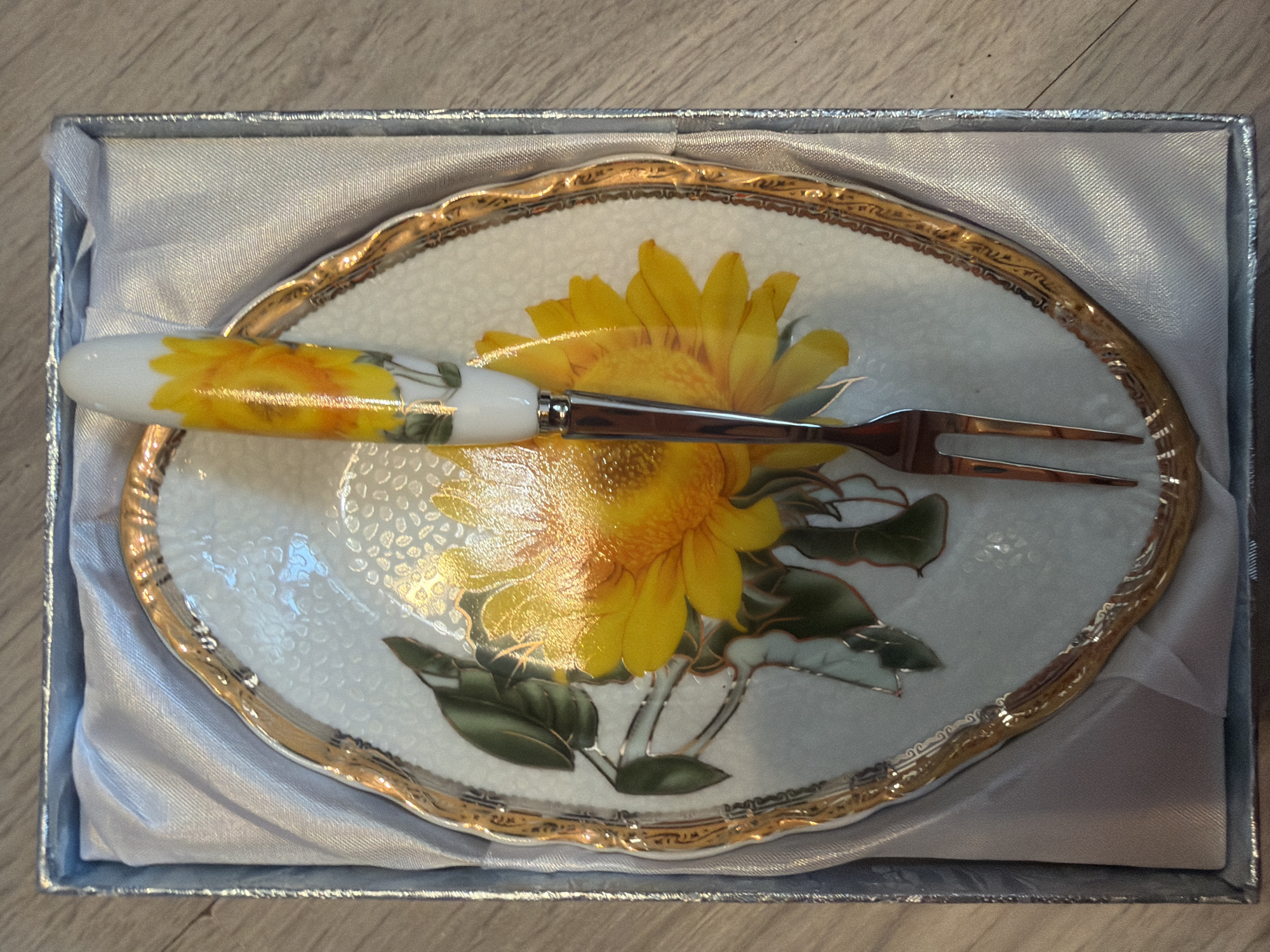 A decorative dessert plate with a matching fork is presented in a silver-lined box. The plate features a large yellow sunflower with green leaves, all rendered in a painted style. The plate has an ornate gold trim around its scalloped edge. The fork has a metallic silver handle and prongs, with a painted sunflower design on the white handle. The box is lined with a shimmering light grey fabric. The background appears to be a light brown wooden surface.