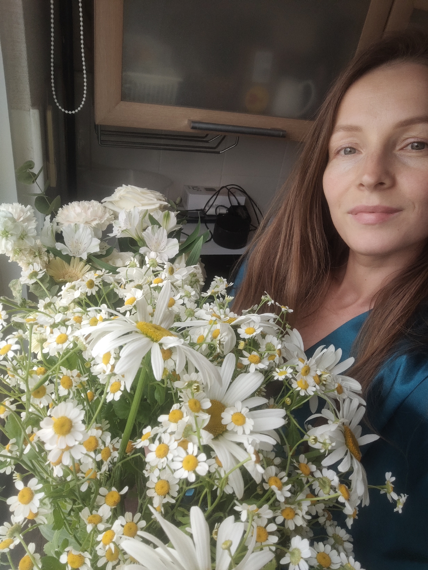 The image depicts a woman holding a large bouquet of white daisies with yellow centers, along with other white flowers. She is wearing a teal-colored top. The background shows a window, cabinets, and some electrical cords. The location is a domestic interior, possibly in the city of Chaikovsky, Russia, though this cannot be confirmed from the image alone.