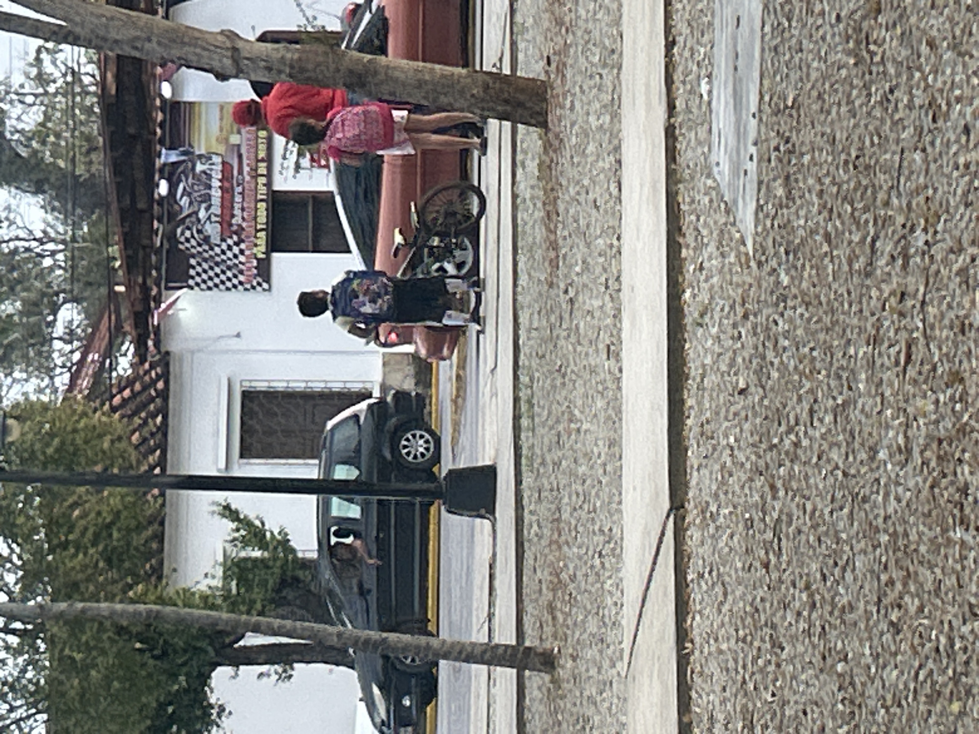 The image shows a street scene in Santa Ana, Venezuela. In the foreground, a rough, gravel-covered area is separated by a concrete curb from a paved street. On the street, a black, four-door hatchback car is parked, with a person partially visible in the driver's seat, leaning out the window. Behind the hatchback, a red, two-door coupe is parked. Next to the coupe, two children, a boy and a girl, are standing. The boy, facing away from the camera, wears a blue backpack and dark shorts with white socks and shoes. The girl, also facing away, wears a pink backpack and white shorts. A woman in a red shirt and red hat is standing next to the girl. In the background, a building with white walls and a terracotta tile roof is visible, with a banner hanging above it that appears to advertise motorcycles and services for "todo tipo de moto" (all types of motorcycles). Trees and telephone poles are also present in the scene. The overall impression is of a casual street encounter on a sunny day.